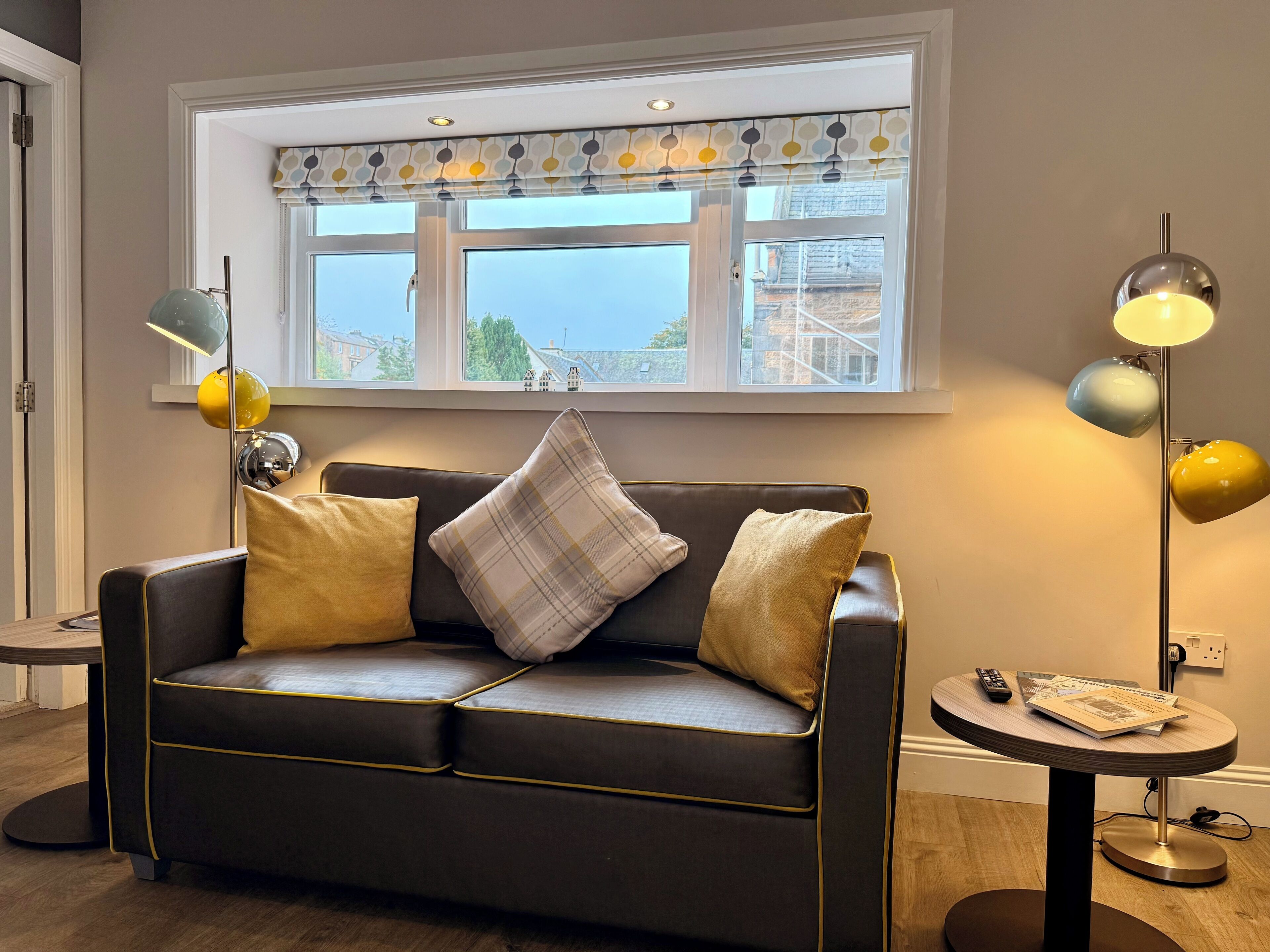A modern living room with a leather sofa, a patterned window valance, and a round side table with a lamp and remote control.