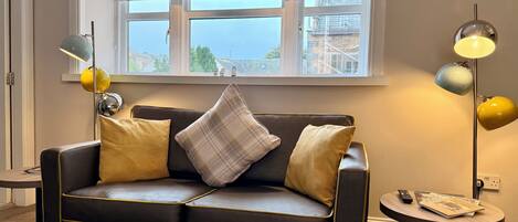 A modern living room with a leather sofa, a patterned window valance, and a round side table with a lamp and remote control.