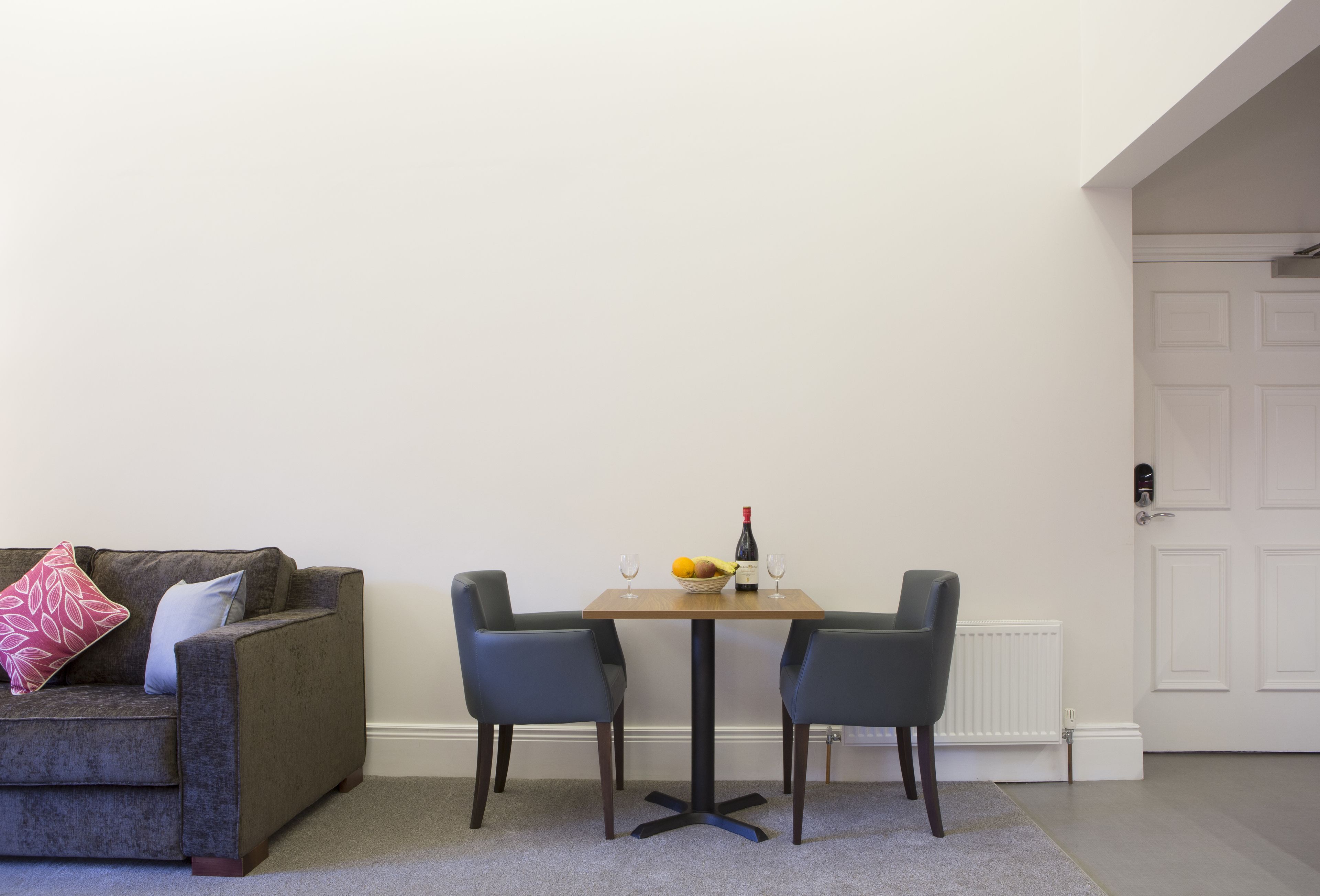A living room with a grey sofa, two armchairs, a small wooden table with a bottle and glasses, and a fruit bowl.
