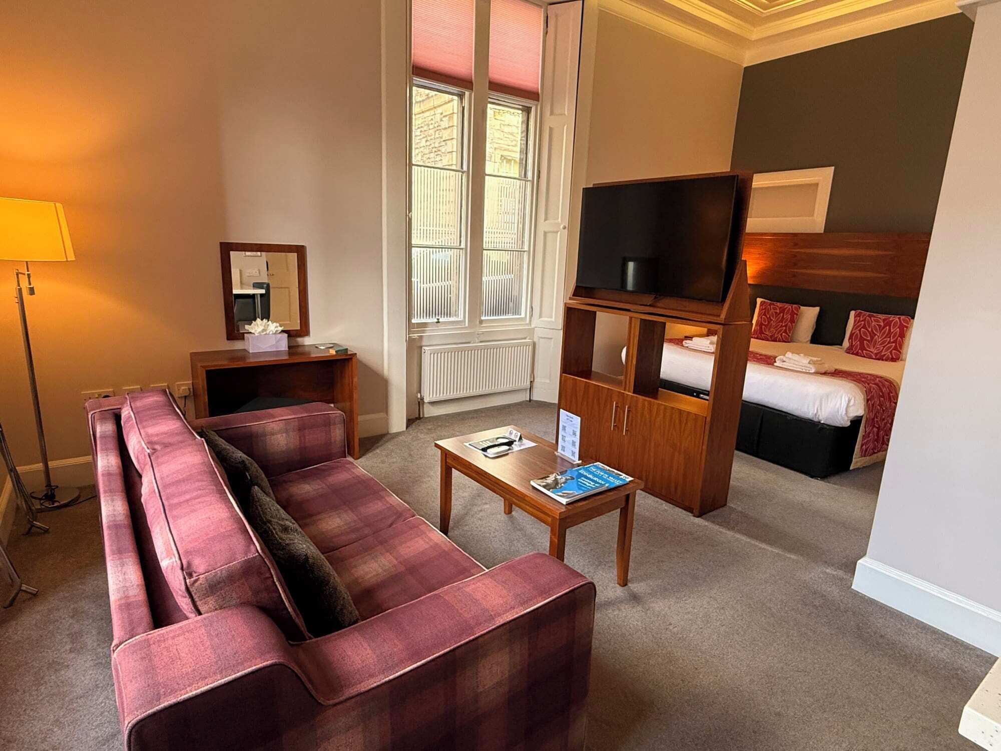 Ein Hotelzimmer mit einer Couch, einem kleinen Tisch, einem Fernseher, einem Bett und einem Fenster mit Jalousien.
