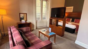 Ein Hotelzimmer mit einer Couch, einem kleinen Tisch, einem Fernseher, einem Bett und einem Fenster mit Jalousien.