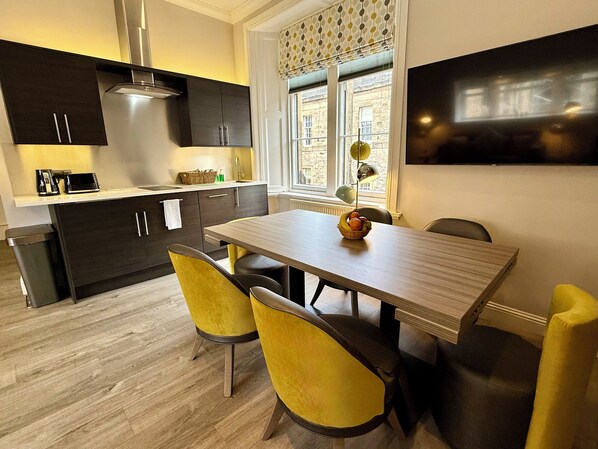 A modern kitchen with dark cabinetry, a stainless steel range hood, and a dining area with a wooden table and yellow chairs.