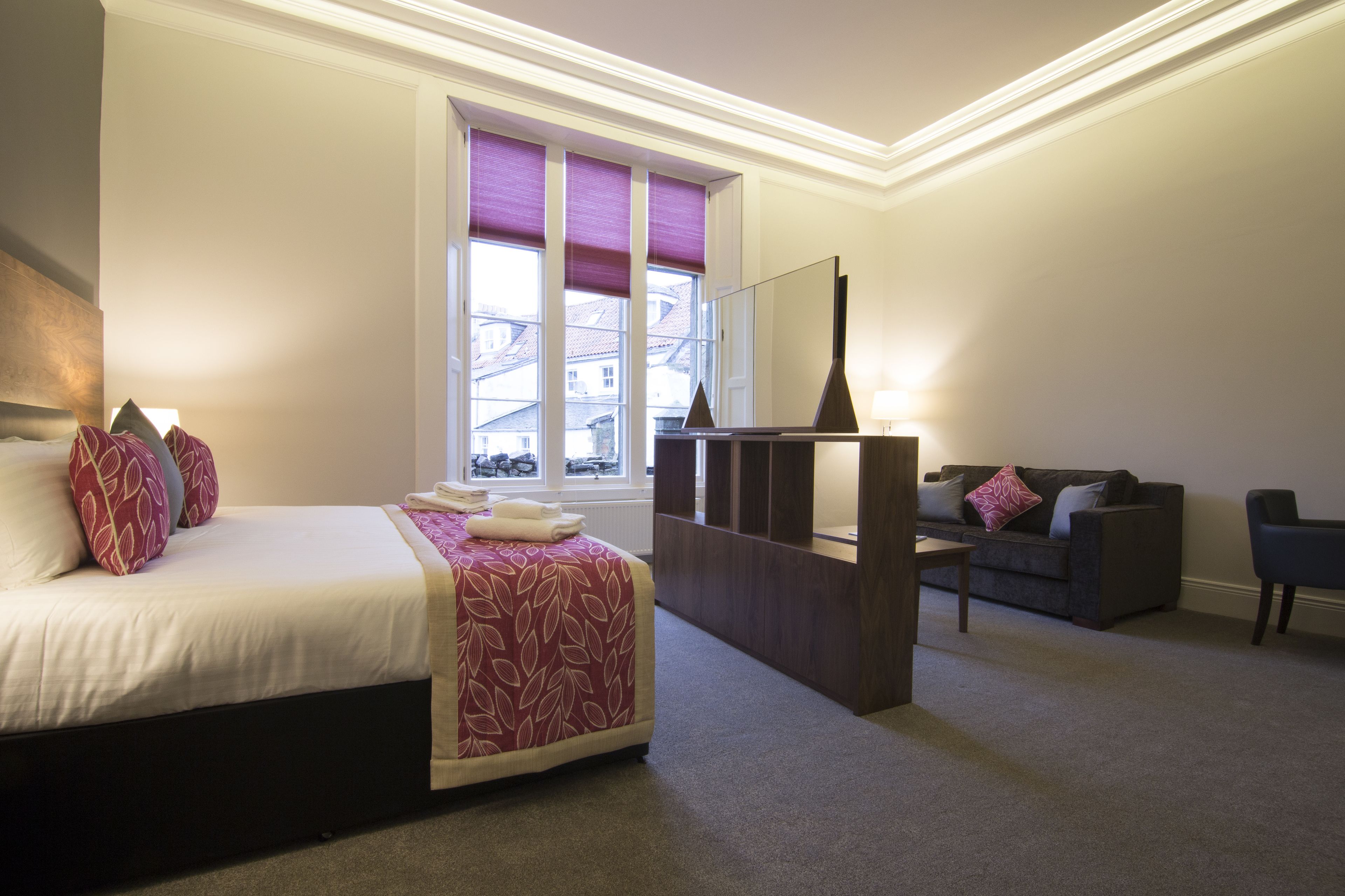 A hotel room with a bed, a sofa, a window with blinds, and a lamp.