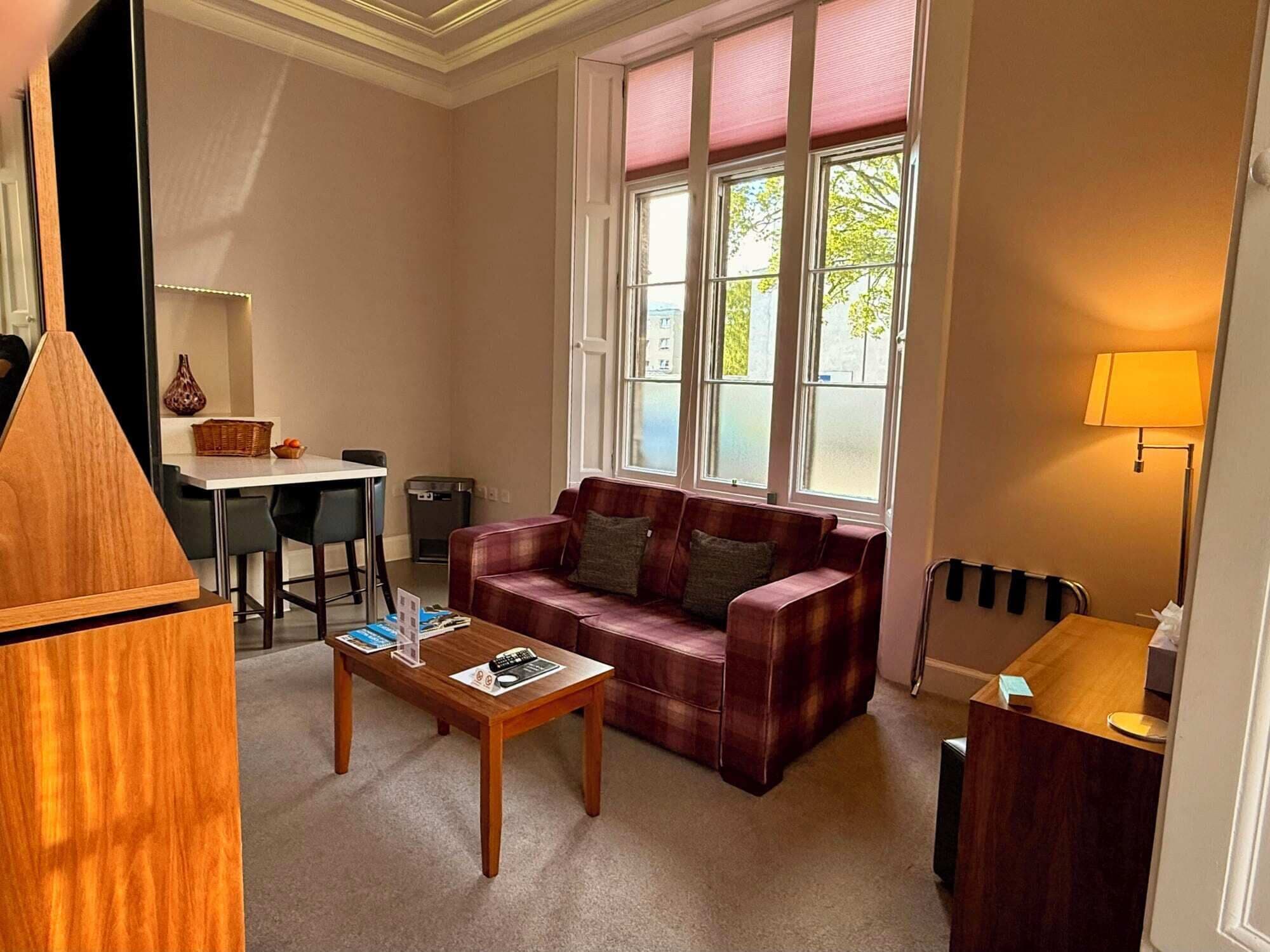 A cozy room with a red sofa, a wooden coffee table, a desk, and a window with blinds.