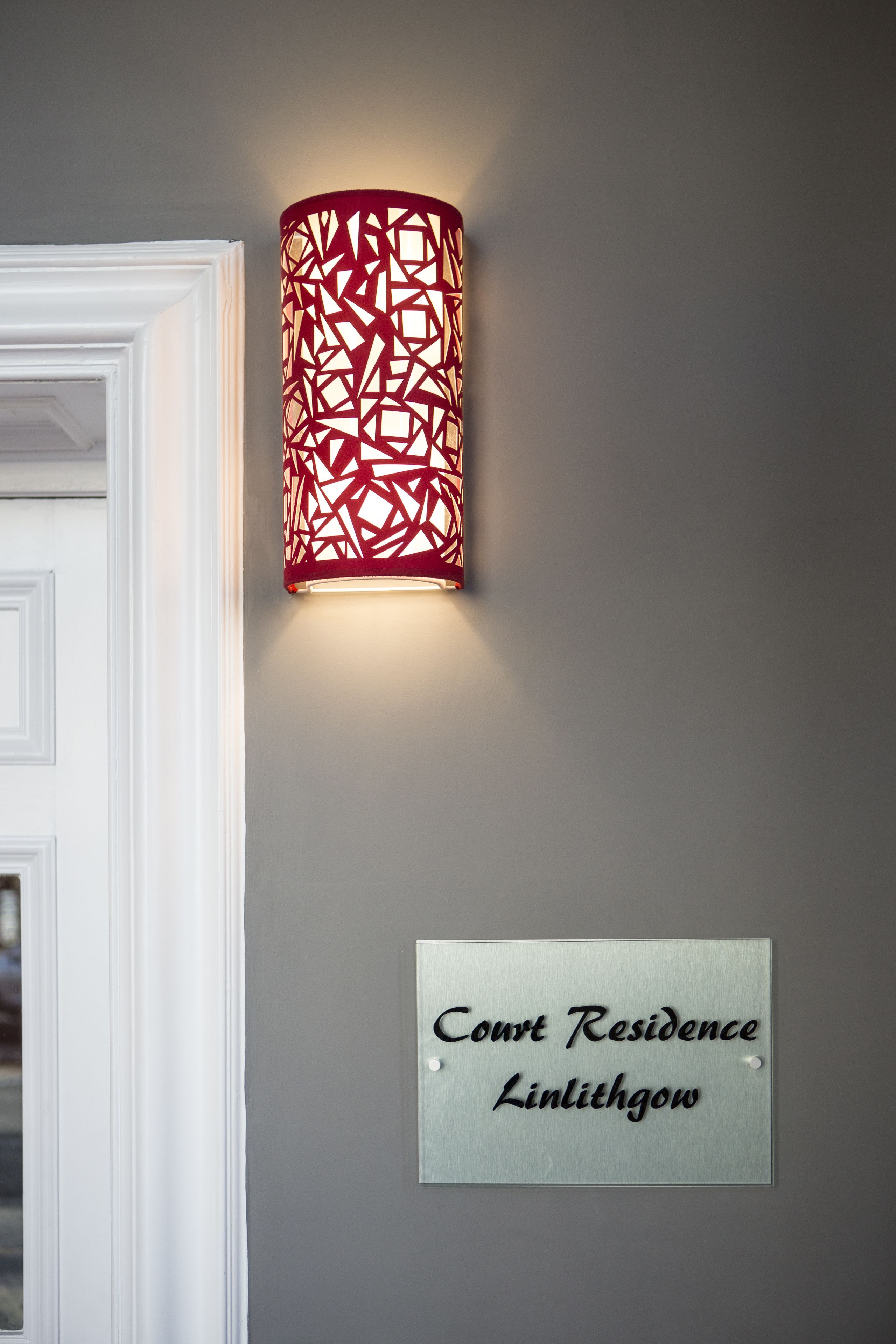 En väggmonterad lampa med ett geometriskt mönster, en dörr med en vit ram och en skylt med texten "Court Residence Linlithgow".