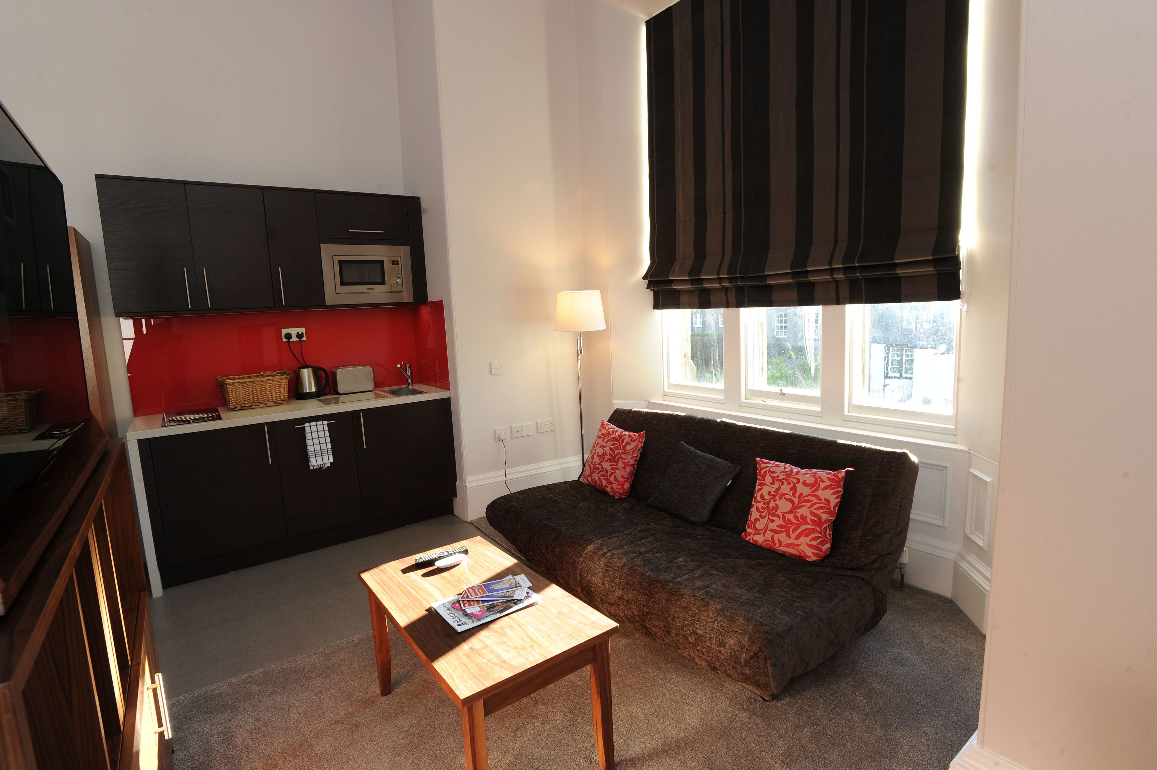 A compact living space with a kitchenette, a sofa with patterned cushions, a small wooden table, and a window with a dark valance.