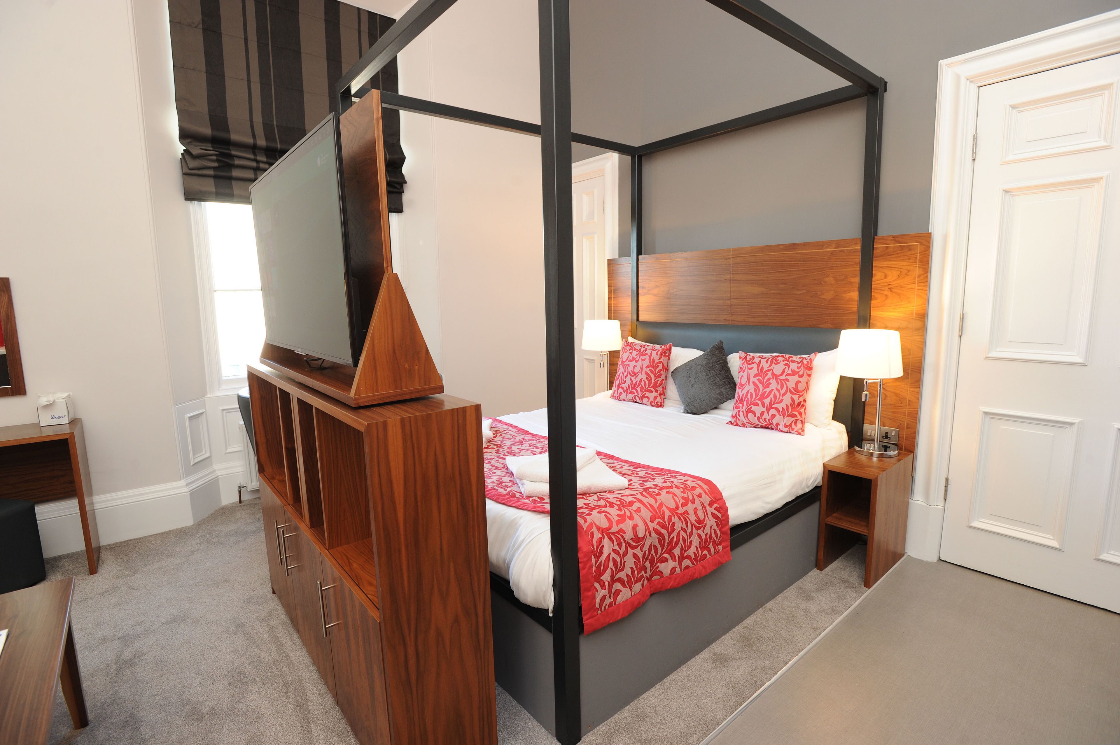 A four-poster canopy bed with a wooden headboard, a bedside table with a lamp, and a TV on a stand.