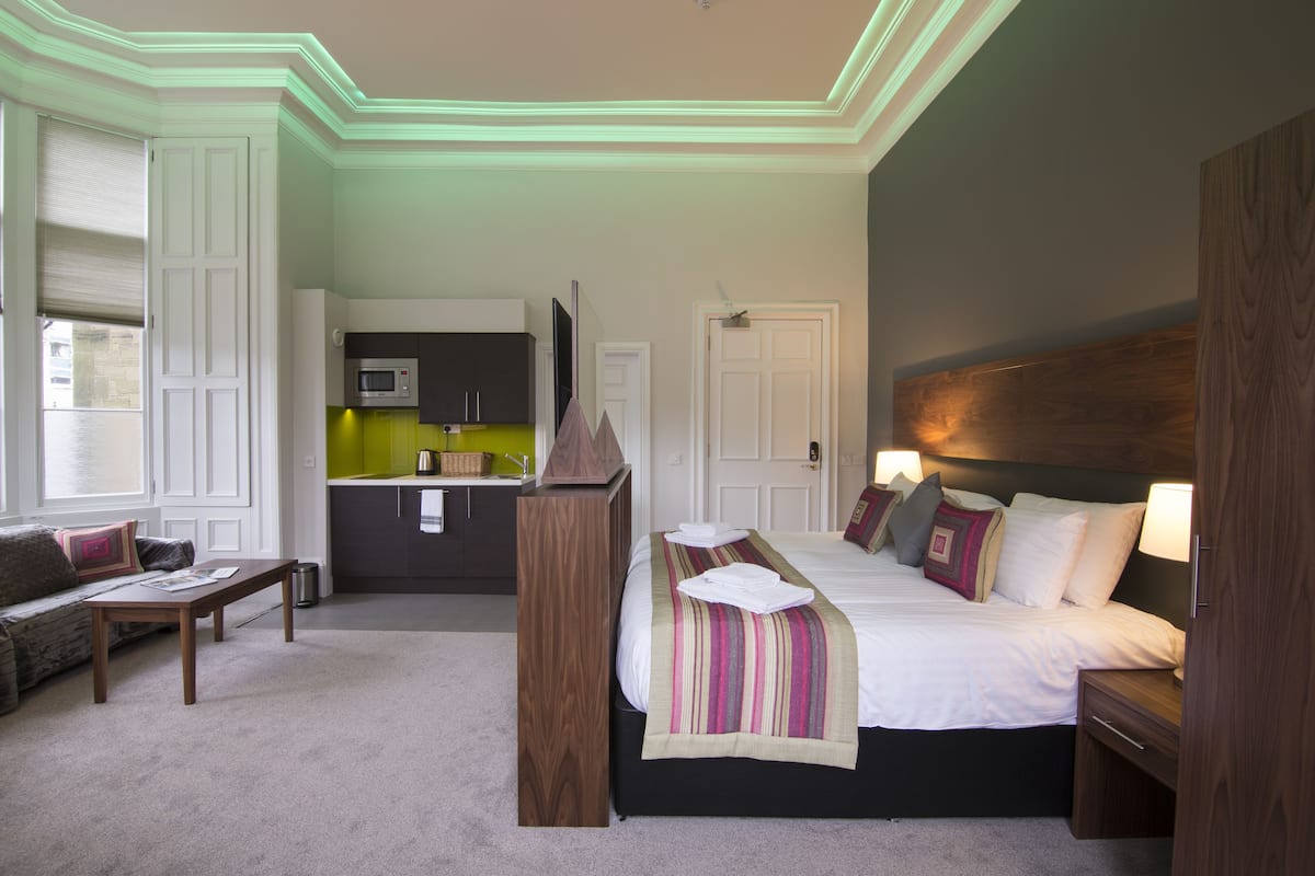 Une chambre à coucher avec un lit, des tables de chevet, une tête de lit en bois, une lampe de chevet, une fenêtre avec des stores, une petite table et, au fond, un coin cuisine.