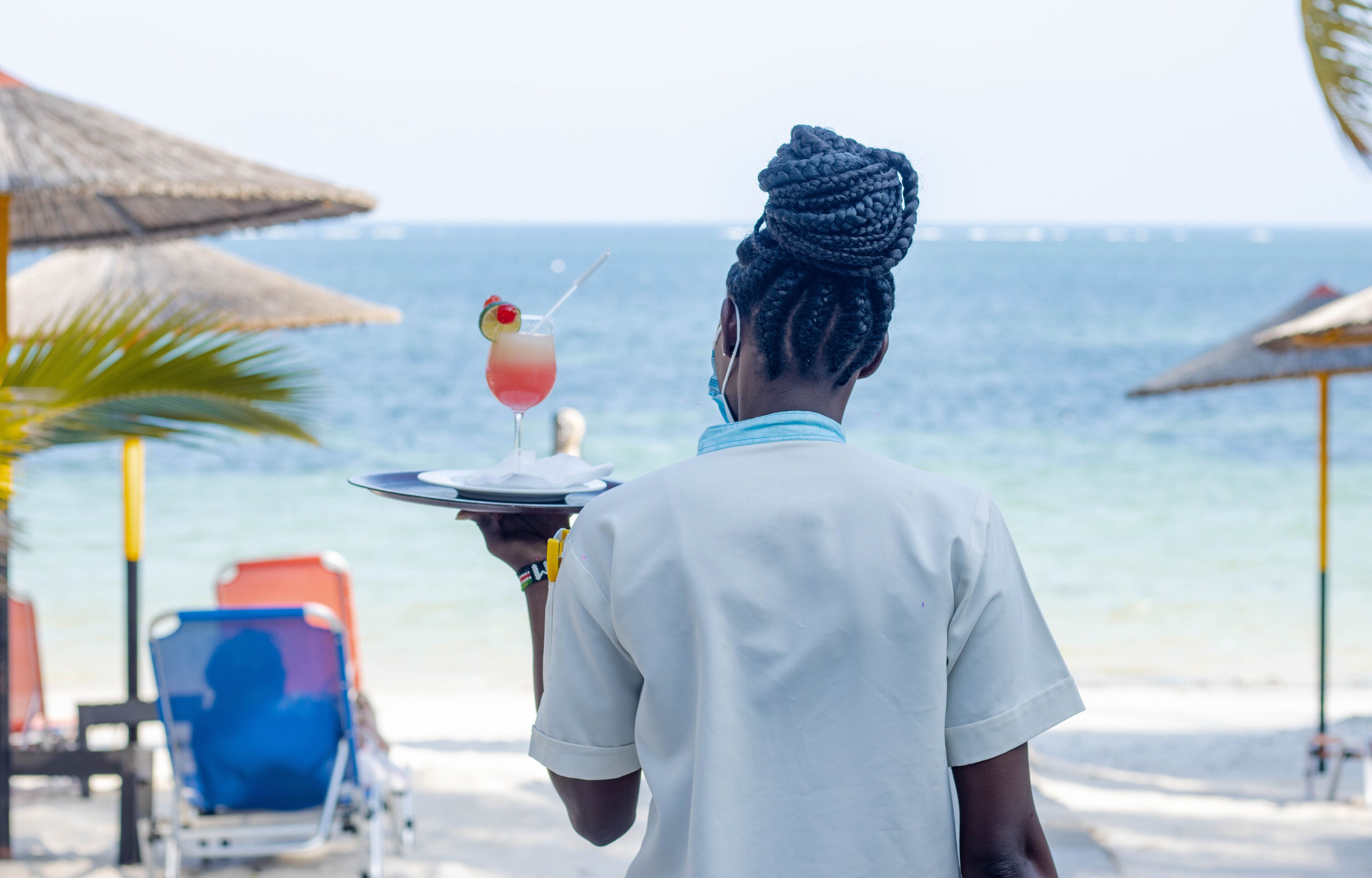 Una persona sosteniendo una bandeja con una bebida y una guarnición de fruta, de pie en una playa con sombrillas y sillas reclinables.