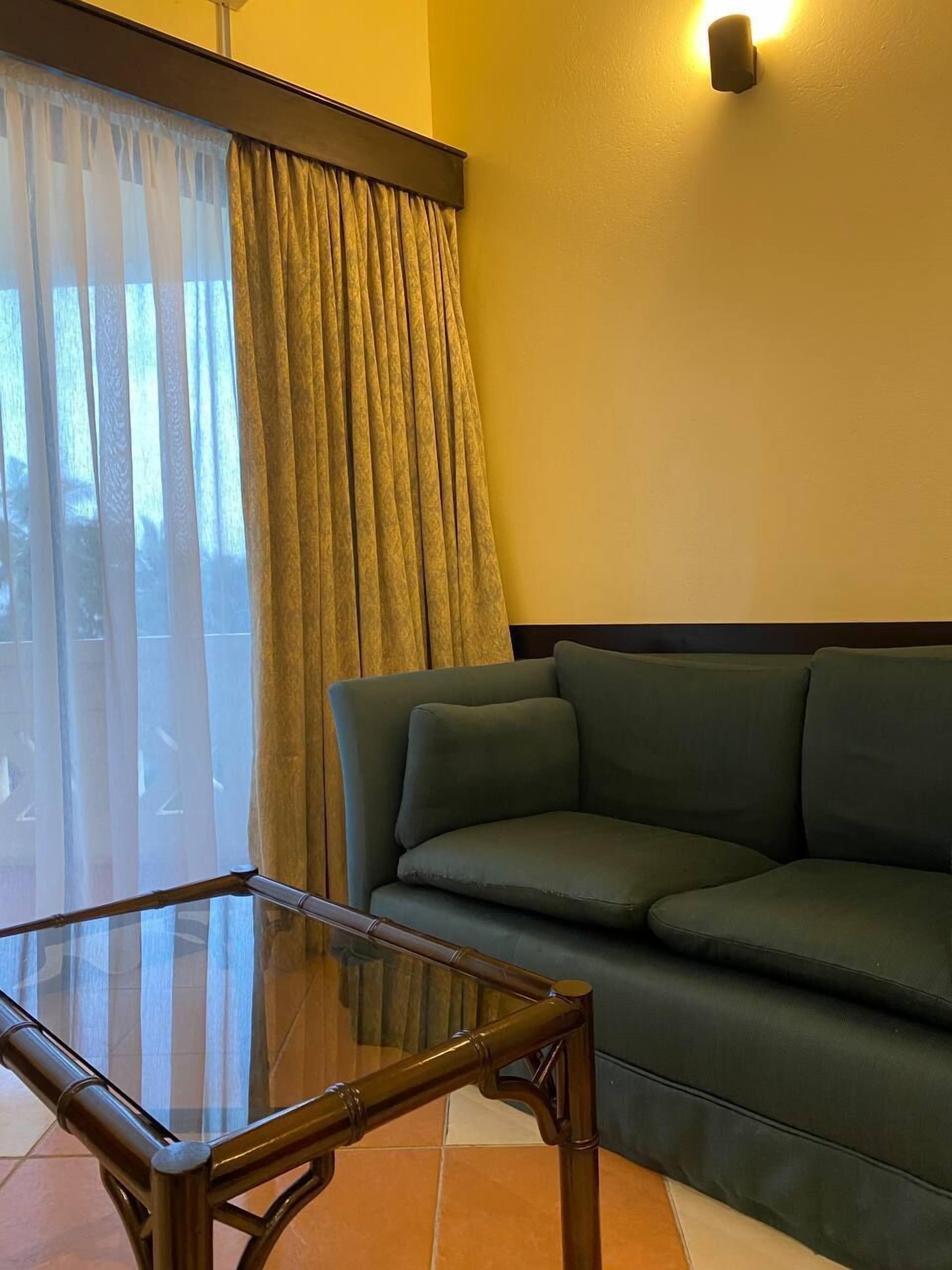 A living room with a glass table, a green sofa, and a large window with curtains.