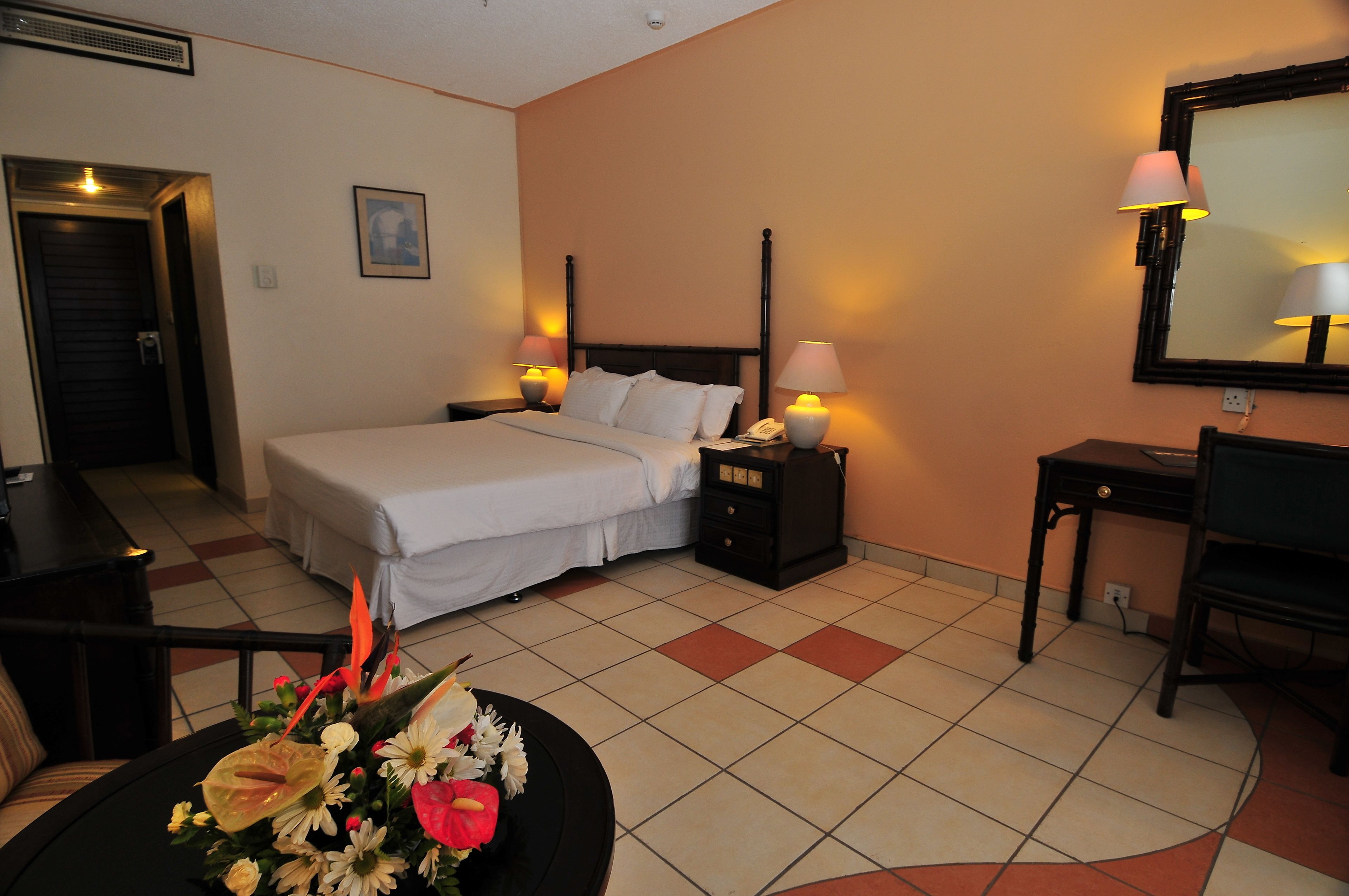 A hotel room with a bed, bedside tables with lamps, a desk, a chair, a mirror, and a flower arrangement.
