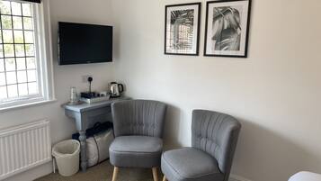 A corner room with two grey upholstered chairs, a flat-screen TV mounted on the wall, a small table with a kettle and a coffee maker, and a window with blinds.