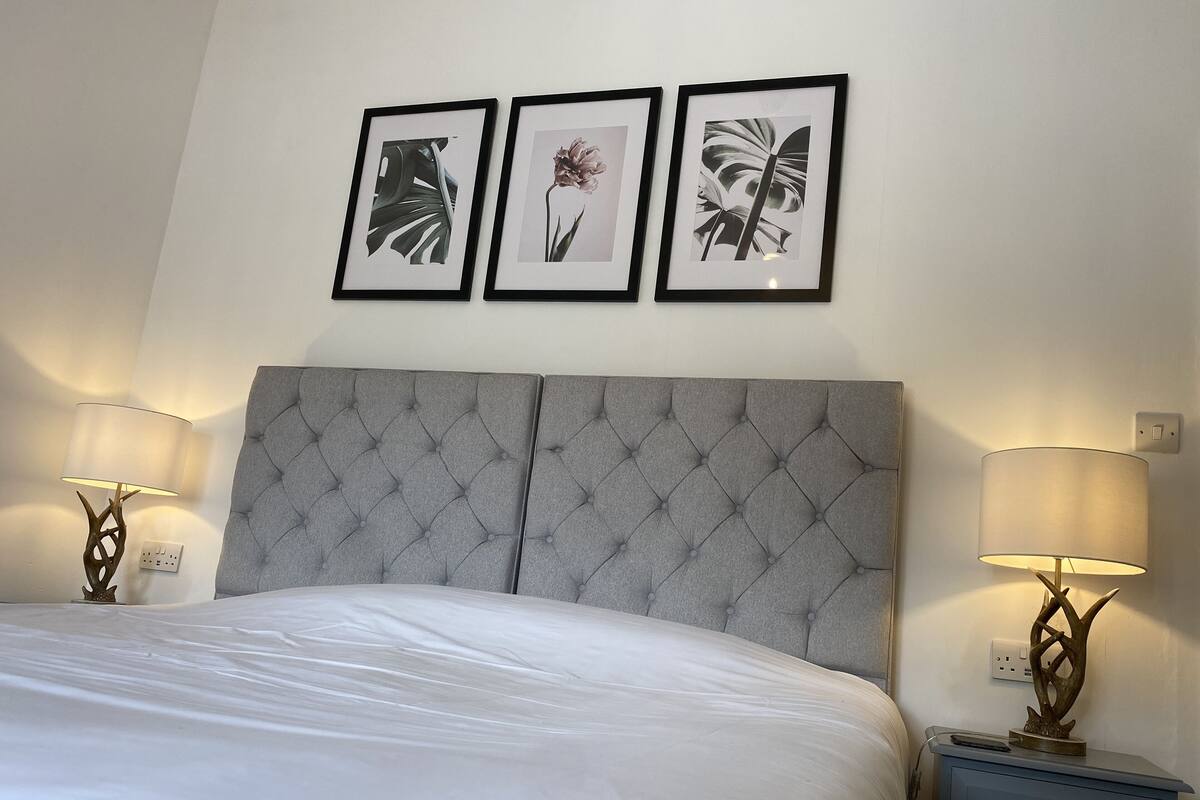 A neatly made bed with a grey tufted headboard, two bedside lamps, and three framed botanical prints on the wall.