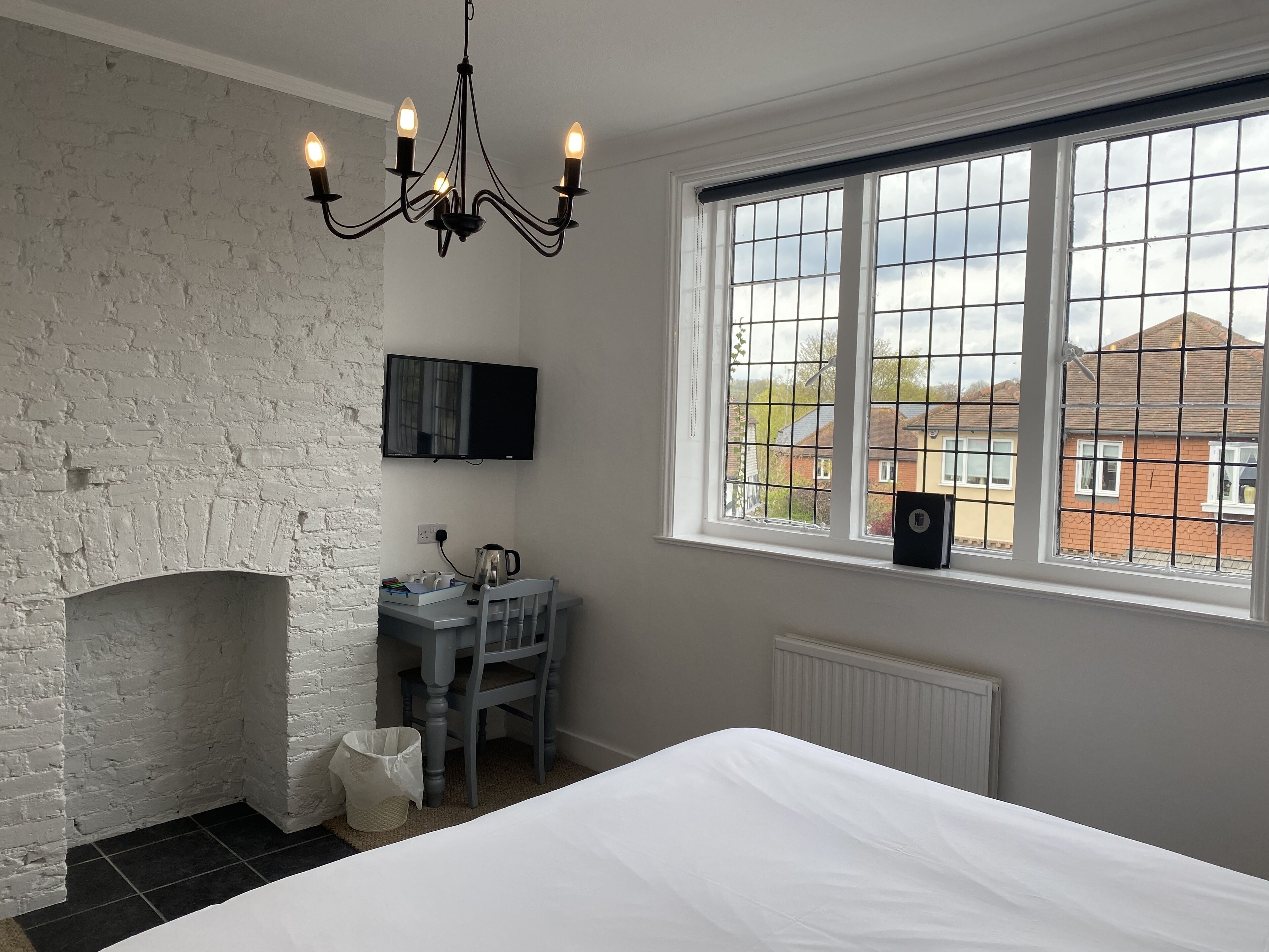 Une chambre avec un mur en briques blanches, une cheminée, un lit, un bureau avec une chaise, un téléviseur et une fenêtre donnant sur des maisons.