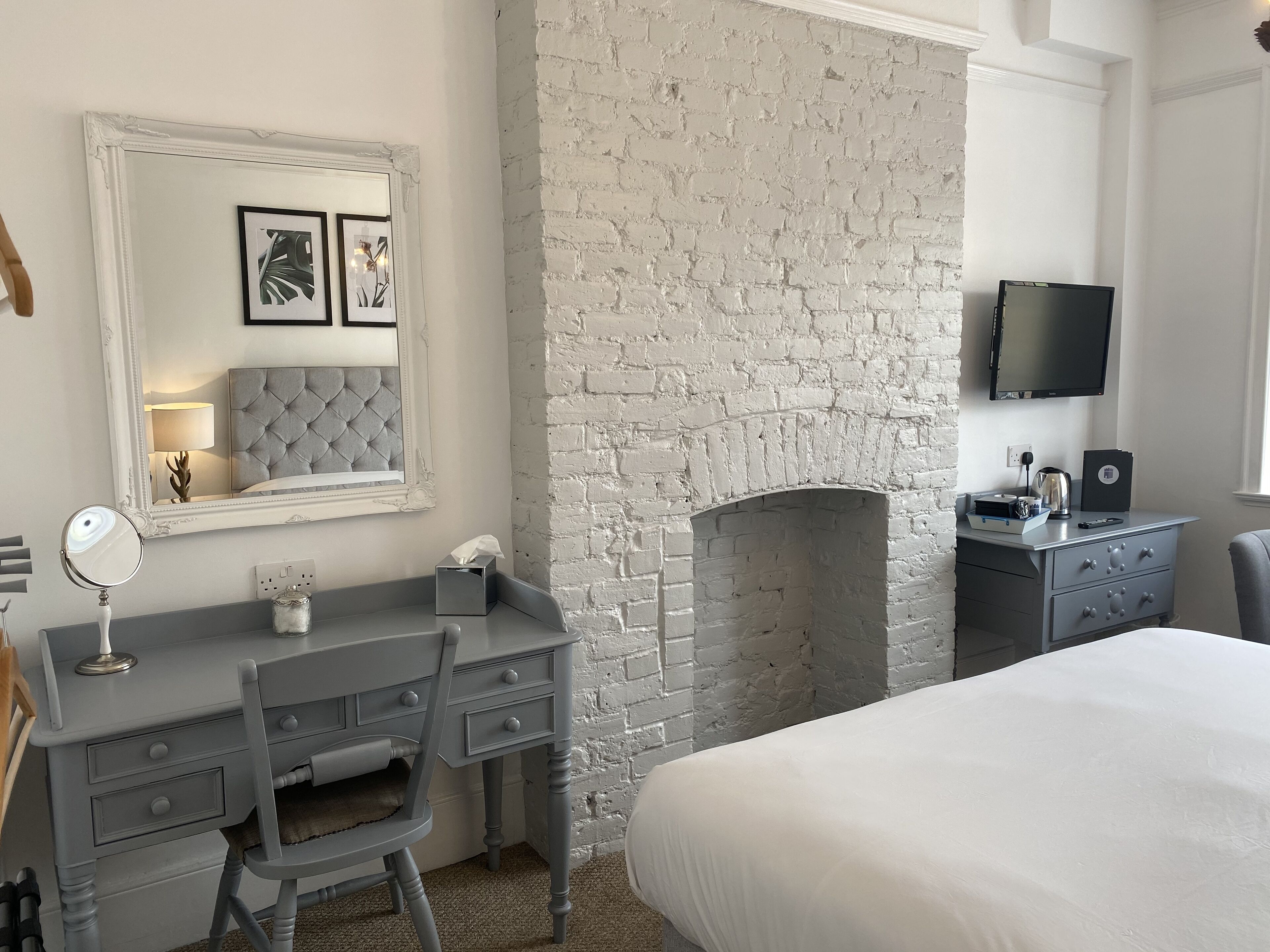 A bedroom with a white brick fireplace, a grey dresser with a mirror, a bed, and a TV mounted on the wall.