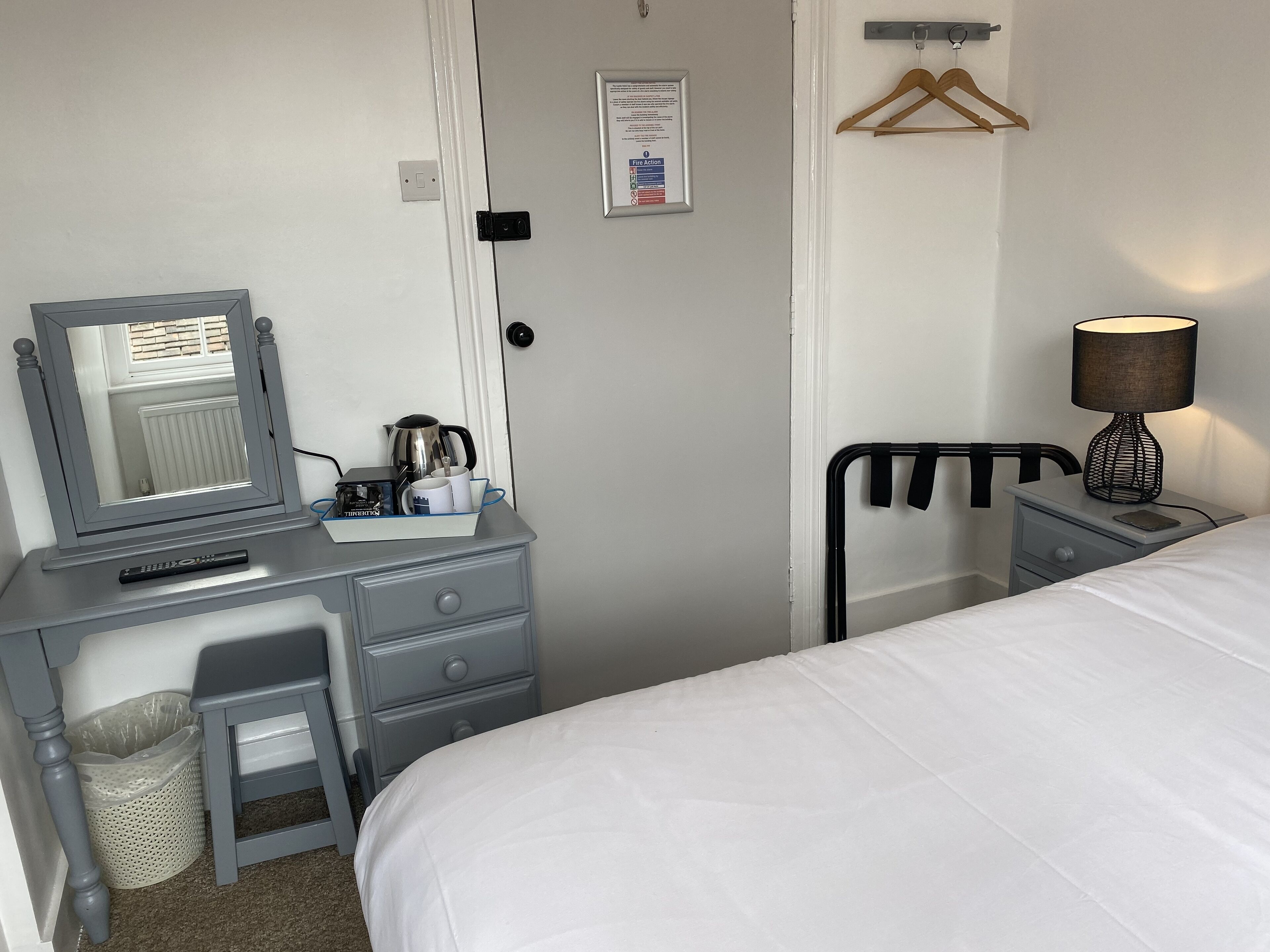 A hotel room with a bed, a desk with a mirror, a lamp, a hanging clothes rack, and a small trash bin.