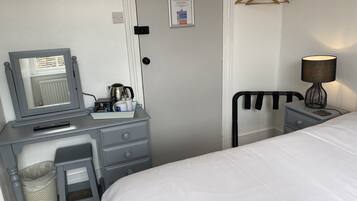 A hotel room with a bed, a desk with a mirror, a lamp, a hanging clothes rack, and a small trash bin.