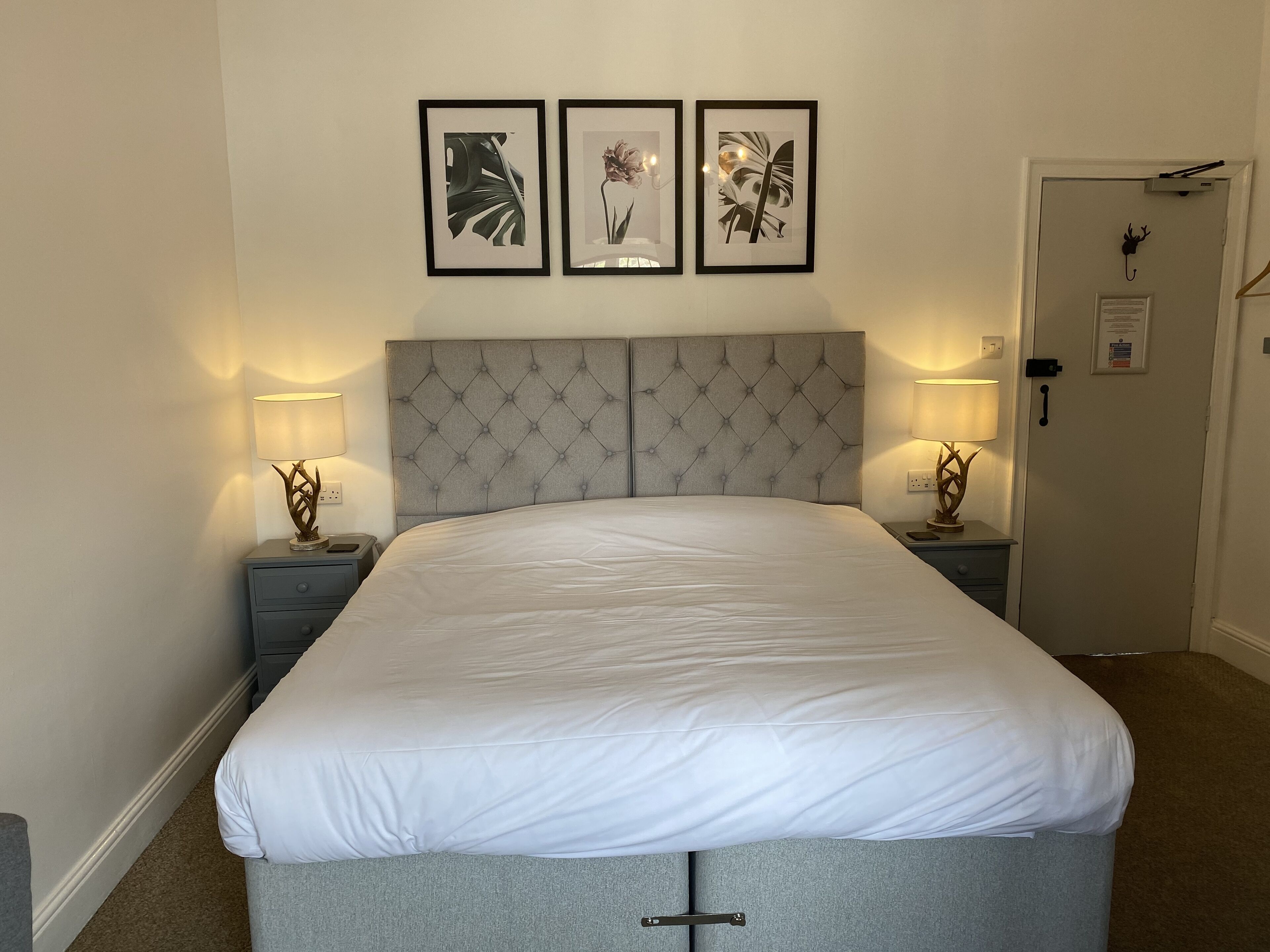 A neatly made bed with a tufted headboard, two bedside tables with lamps, and three framed botanical prints on the wall.
