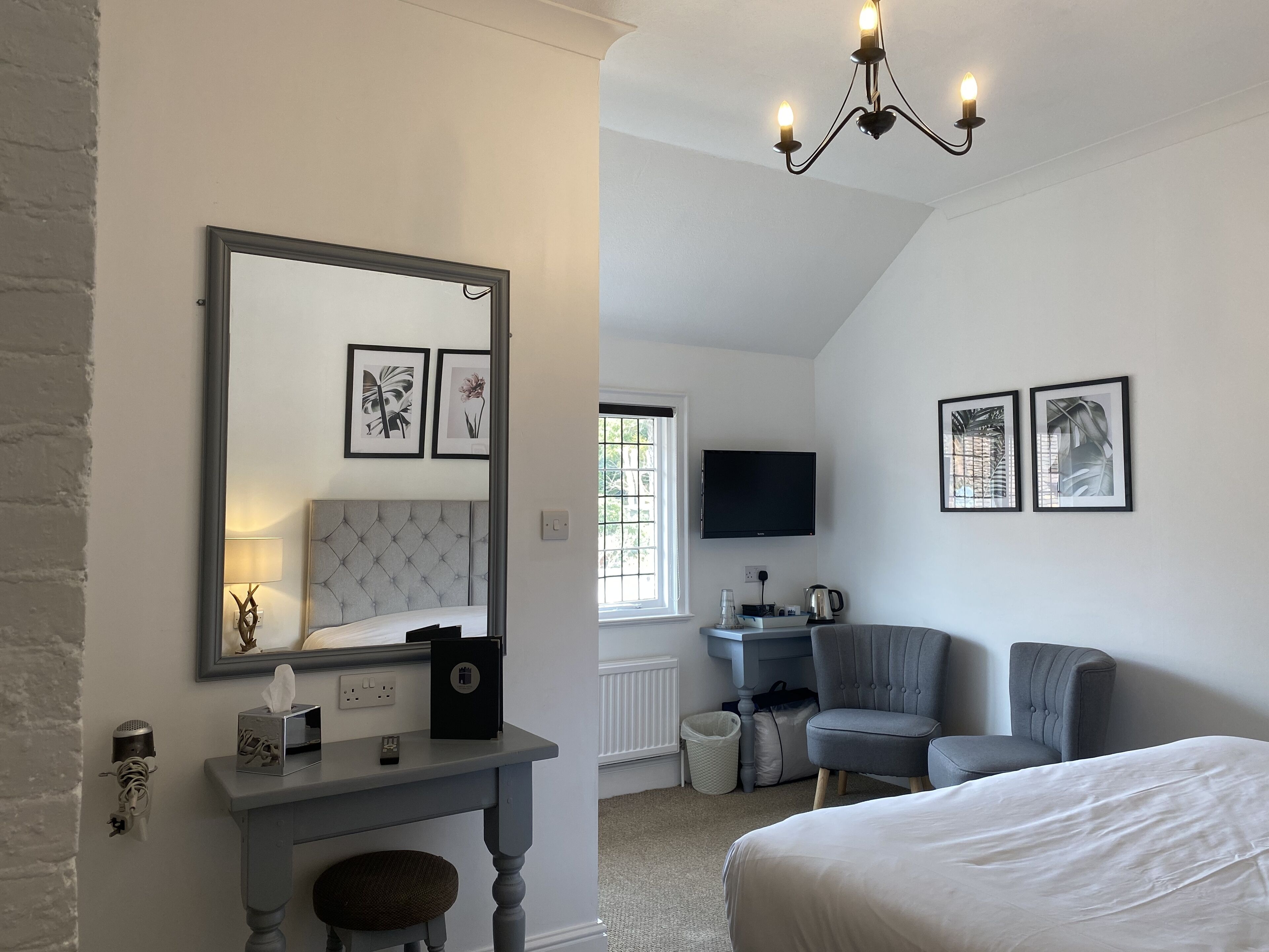 A bedroom with a bed, a vanity with a mirror, a desk, a chair, a television, and artwork on the walls.