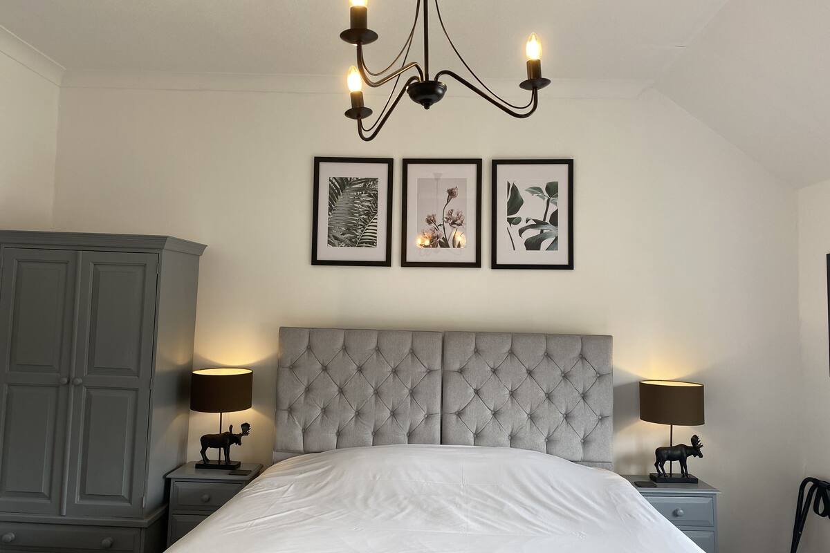 A bedroom with a tufted headboard, a bed with white bedding, two bedside tables with lamps, and three framed botanical prints on the wall.