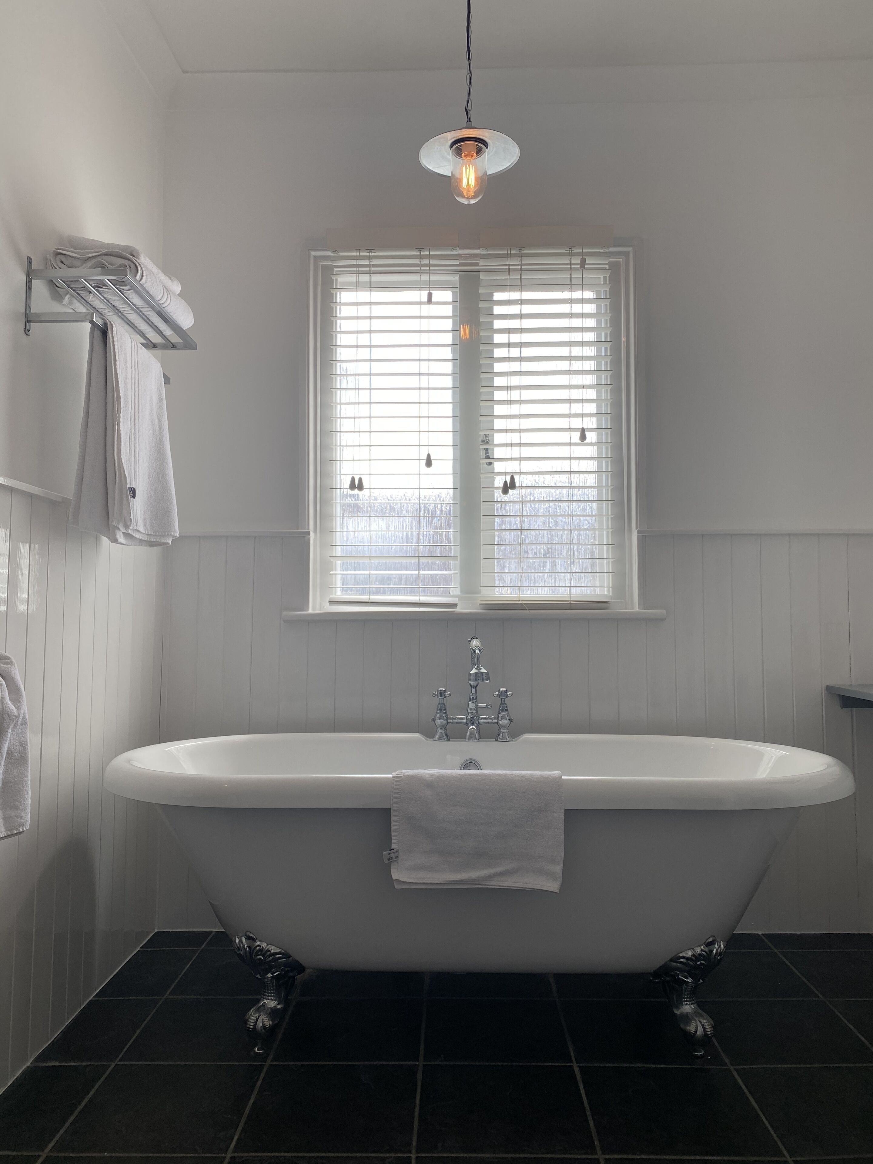 A bathroom with a freestanding bathtub, white walls, a window with blinds, and a hanging light fixture.