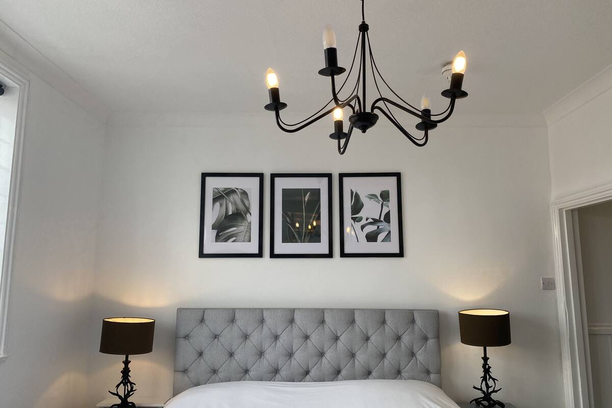 A bedroom with a tufted headboard, a chandelier, two bedside tables with lamps, and three framed botanical prints on the wall.