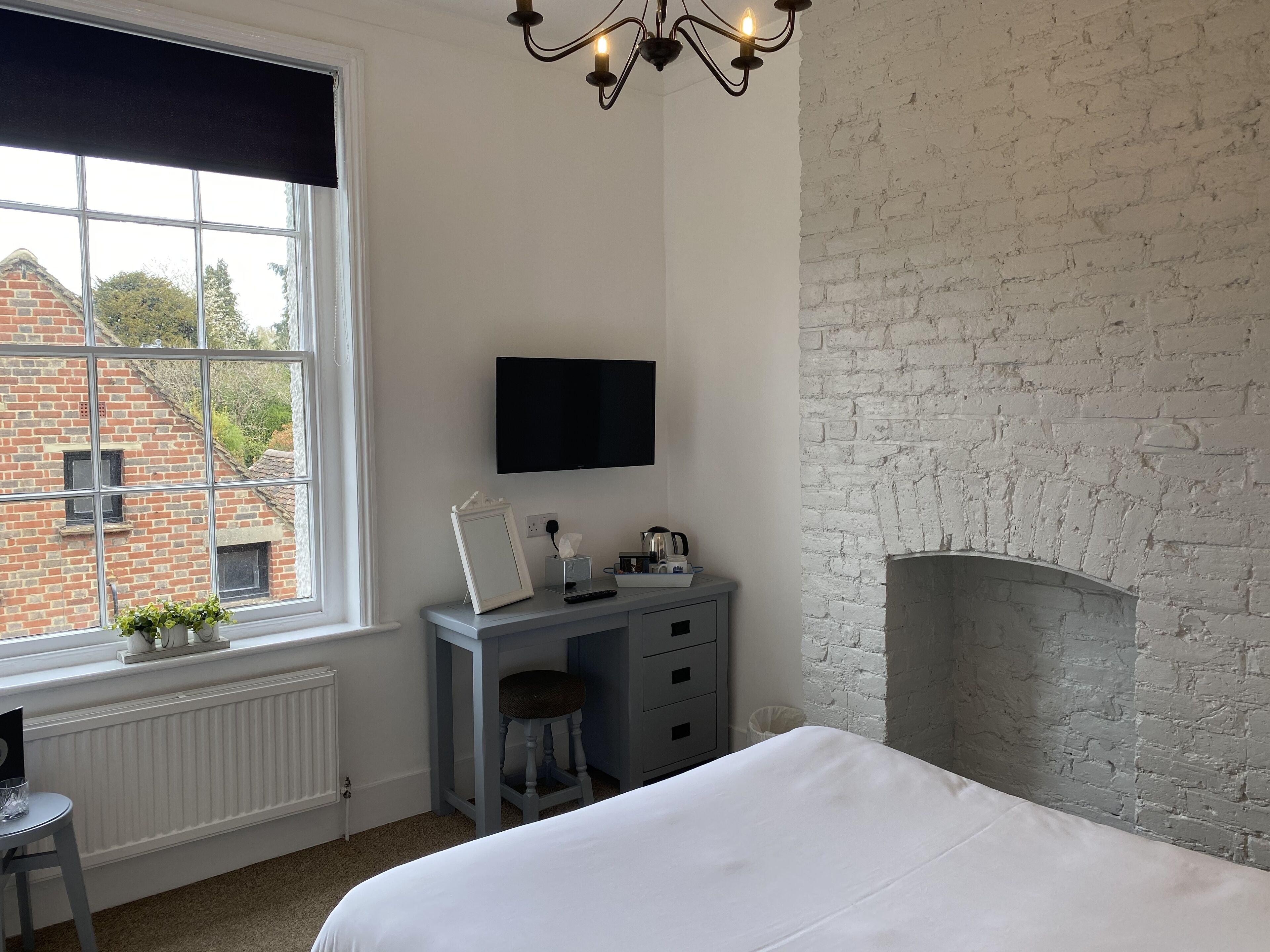 A bedroom with a large window, a desk, a chair, a bed, a flat-screen TV, and a fireplace.