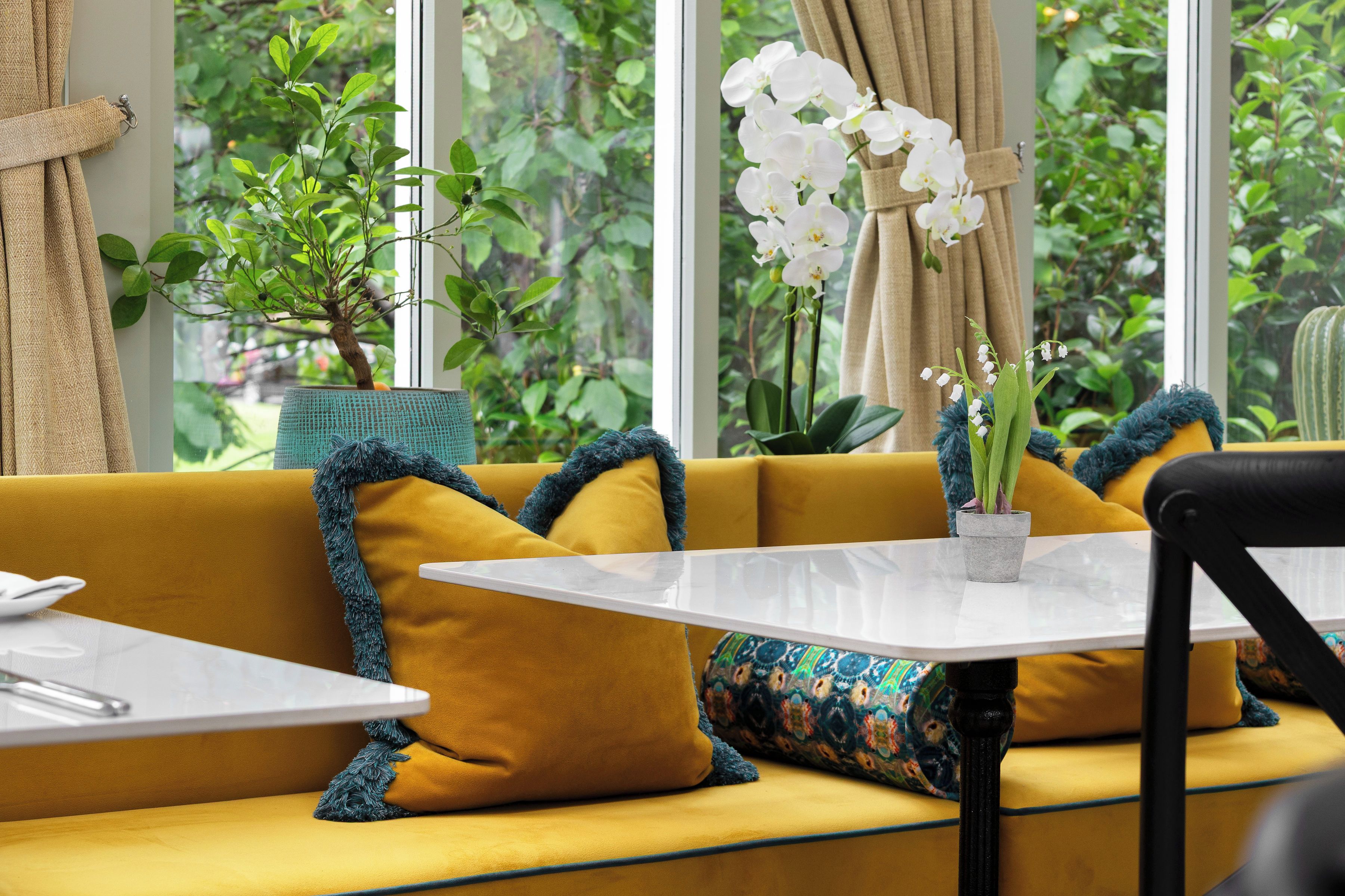 Un canapé jaune, une table blanche et une plante devant une fenêtre donnant sur un paysage verdoyant.