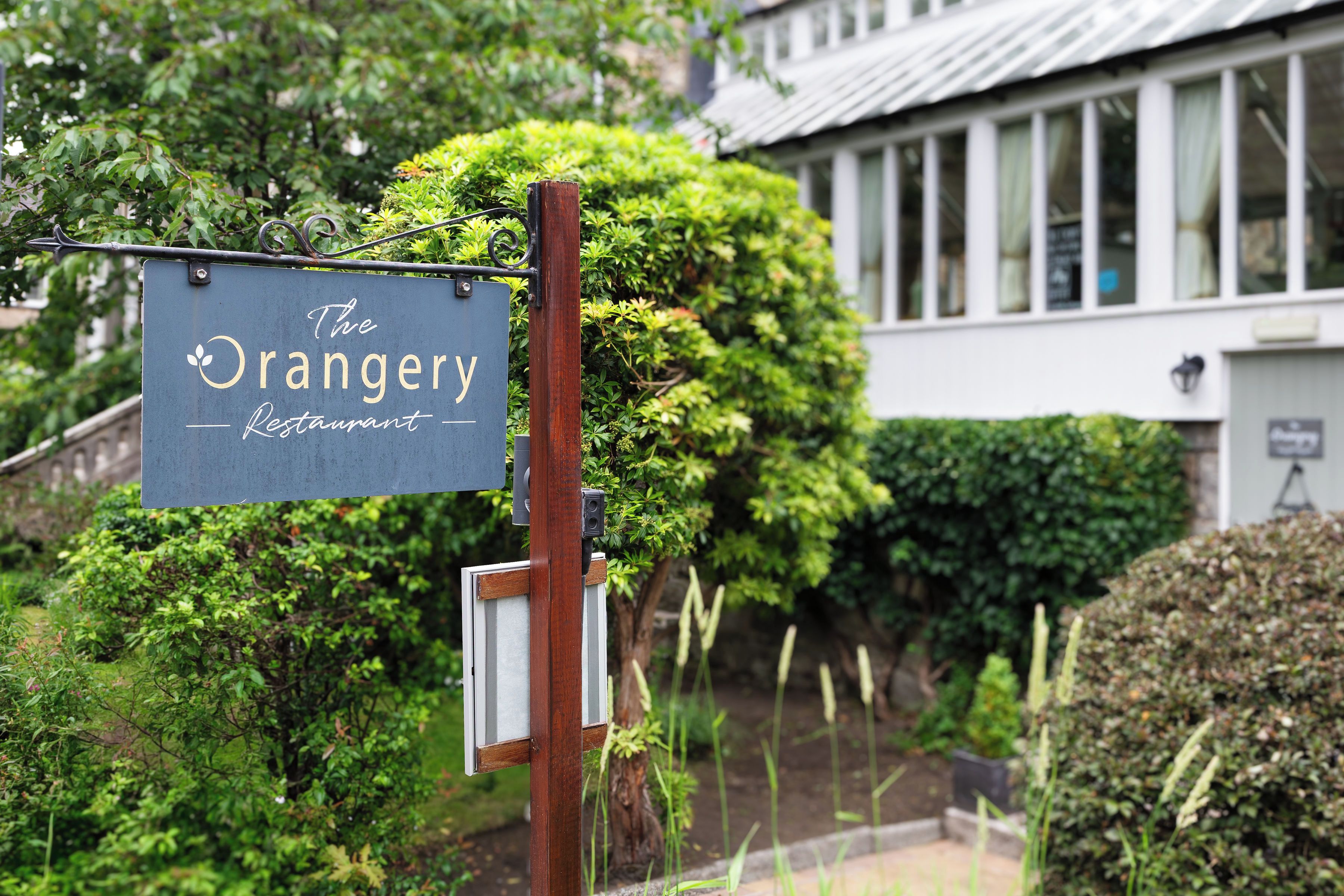A sign for 'The Orangery Restaurant' mounted on a metal post with a wooden post nearby, set against a backdrop of lush greenery and a building with large windows.