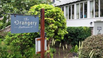 A sign for 'The Orangery Restaurant' mounted on a metal post with a wooden post nearby, set against a backdrop of lush greenery and a building with large windows.
