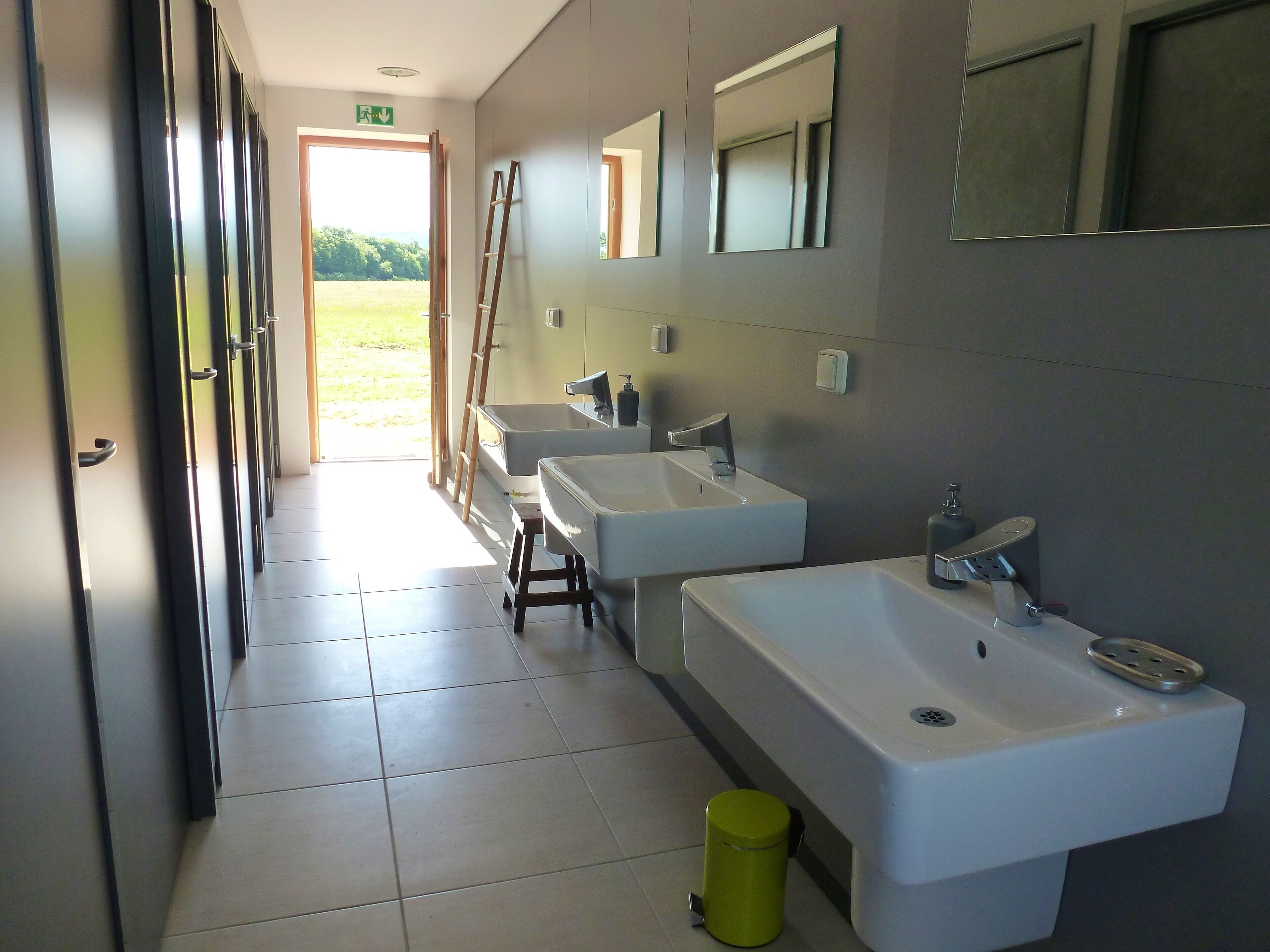 Un baño moderno con dos lavabos, un espejo y un inodoro. Una escalera está apoyada en la pared cerca de la puerta.