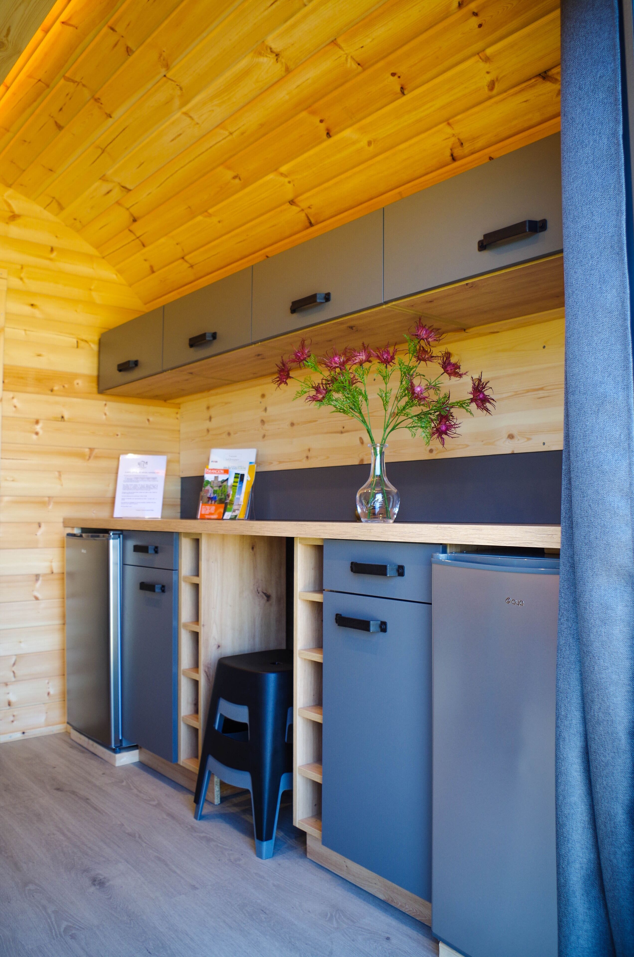A compact kitchen with wooden cabinets, a black stool, and a refrigerator.