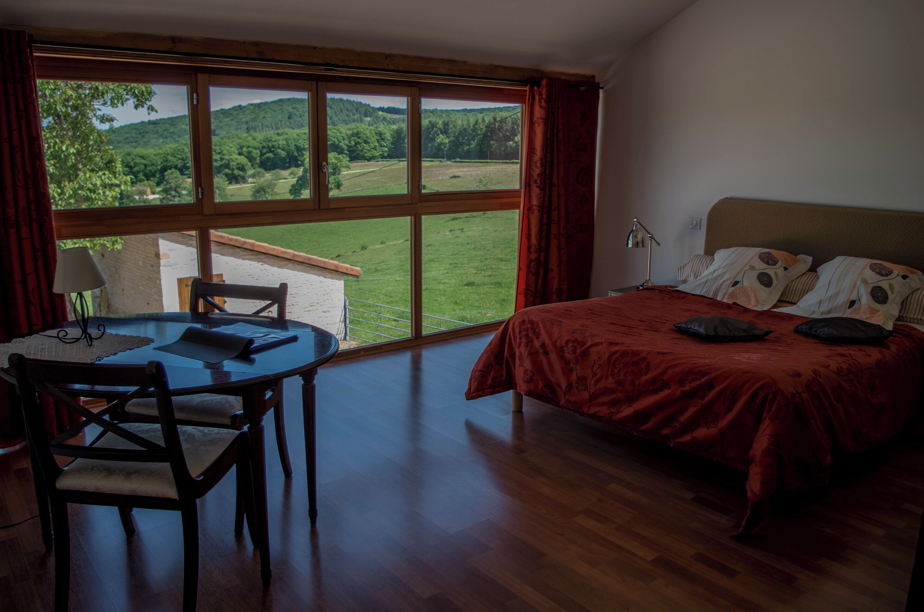 Ein Schlafzimmer mit einem großen Fenster, einem Bett mit roter Bettdecke und einem Holztisch mit Stühlen.