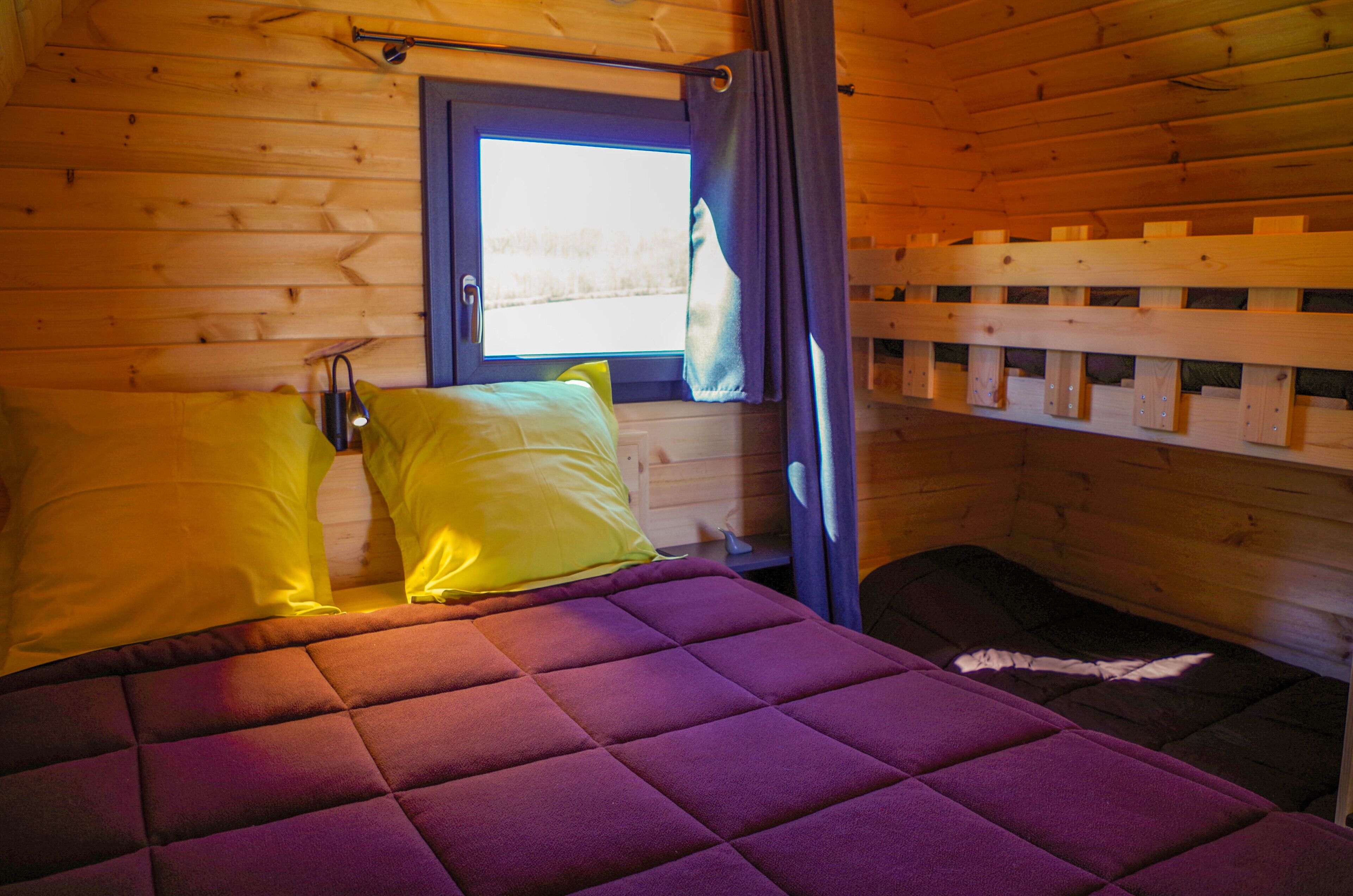 Una habitación de cabaña de madera con una cama, un ventanal con cortinas y una litera.