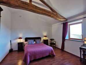 Une chambre avec un plafond en bois, un lit recouvert d’une couverture violette, deux lampes de chevet, une chaise et une fenêtre avec des rideaux.