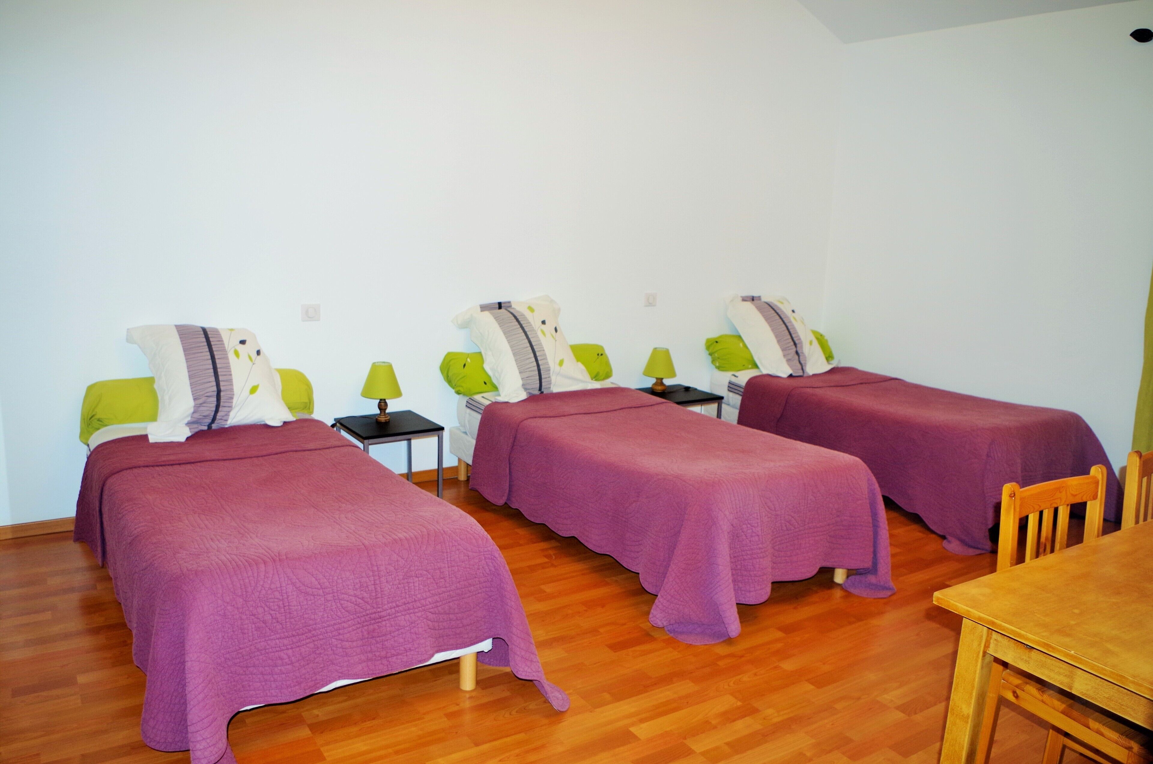 A room with three beds, each with a purple bedspread and green pillows, a small table, and a wooden chair.