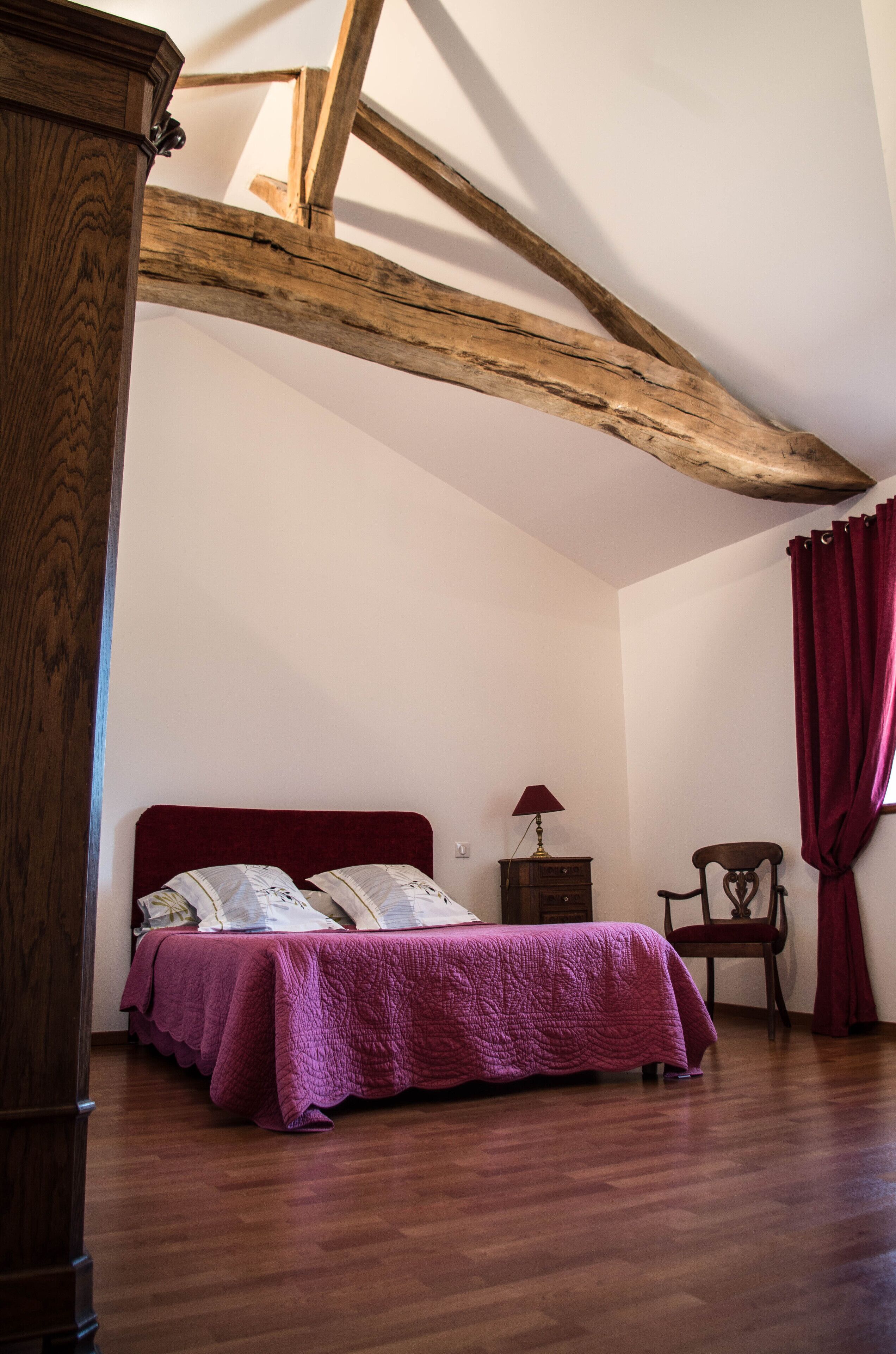 Une chambre avec un plafond en bois, un lit recouvert d’une couverture rouge, une table de chevet, une chaise et un rideau rouge.