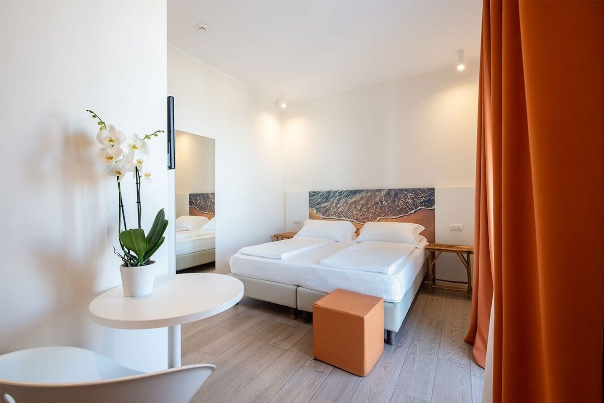 Une chambre d’hôtel moderne avec un grand lit, une petite table ronde, une plante en pot et une vue sur le paysage urbain par la fenêtre.