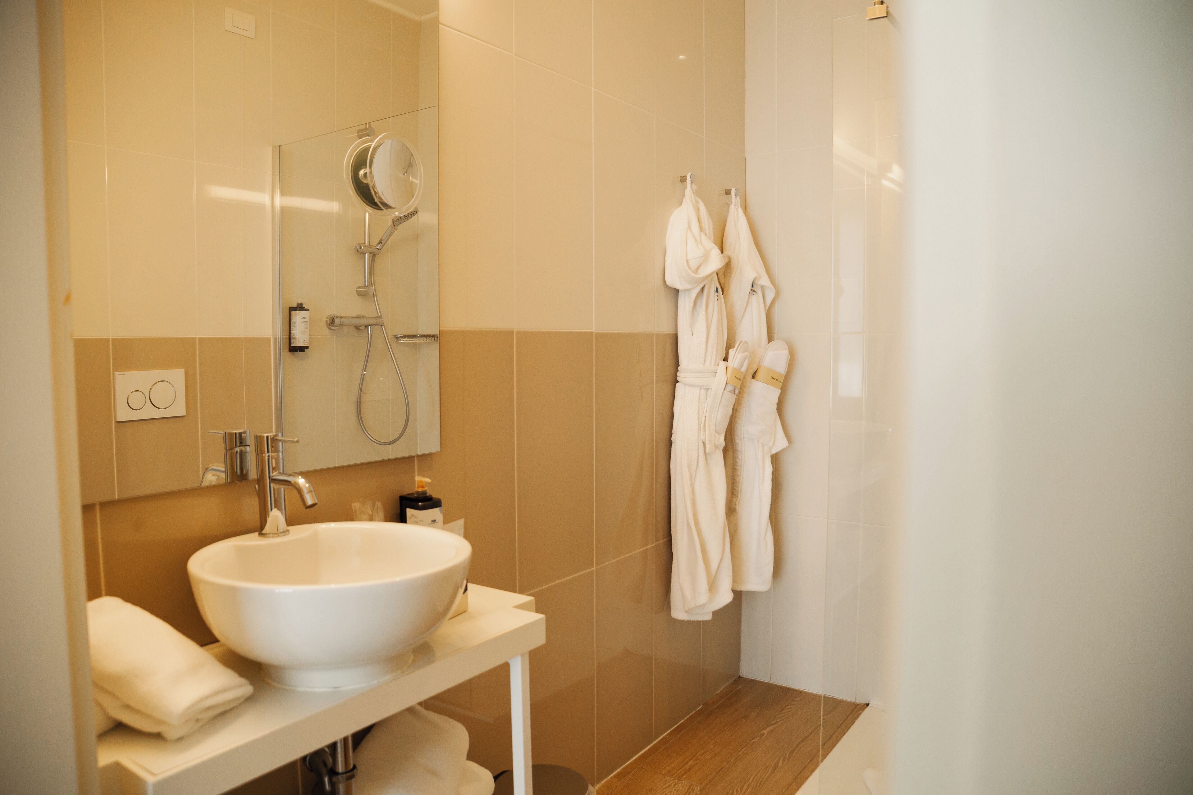 Une salle de bain moderne avec un grand lavabo blanc, un miroir, une douche et deux peignoirs blancs suspendus à des crochets.