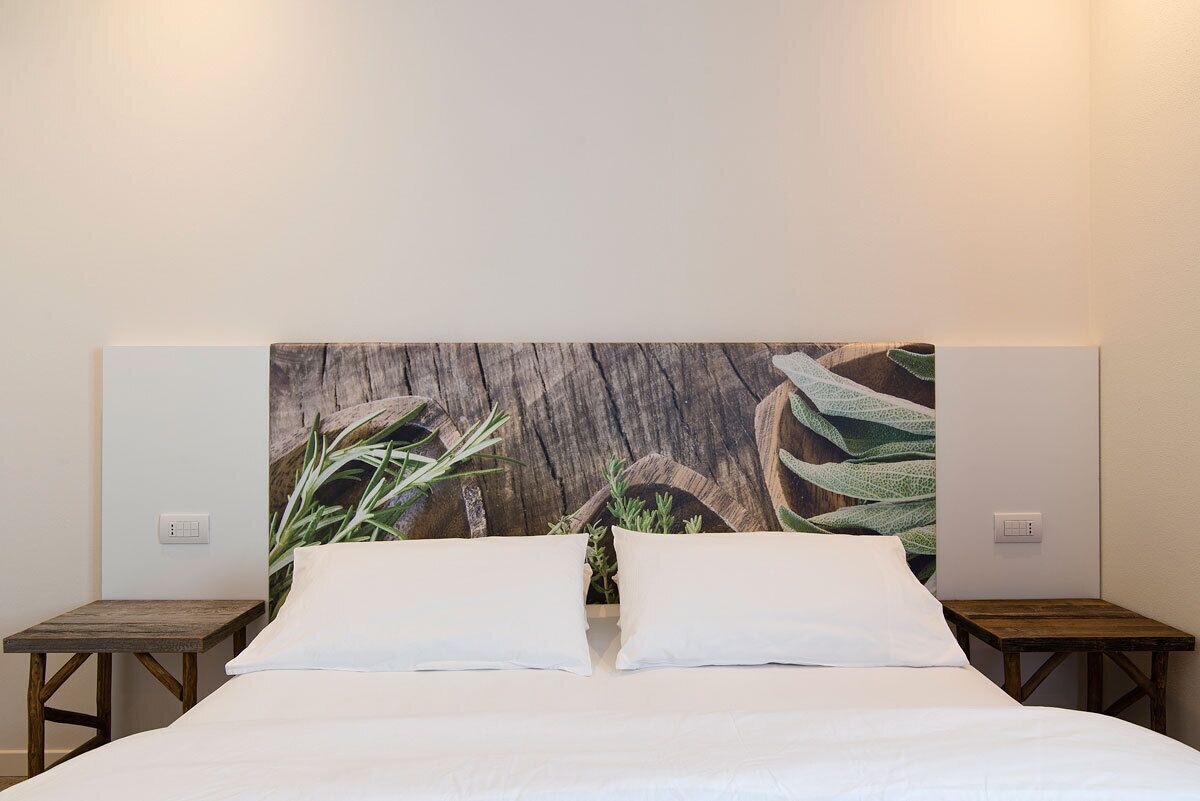 Un lit recouvert de draps blancs, flanqué de deux tables de chevet en bois, adossé à un mur orné d’un grand tableau abstrait.