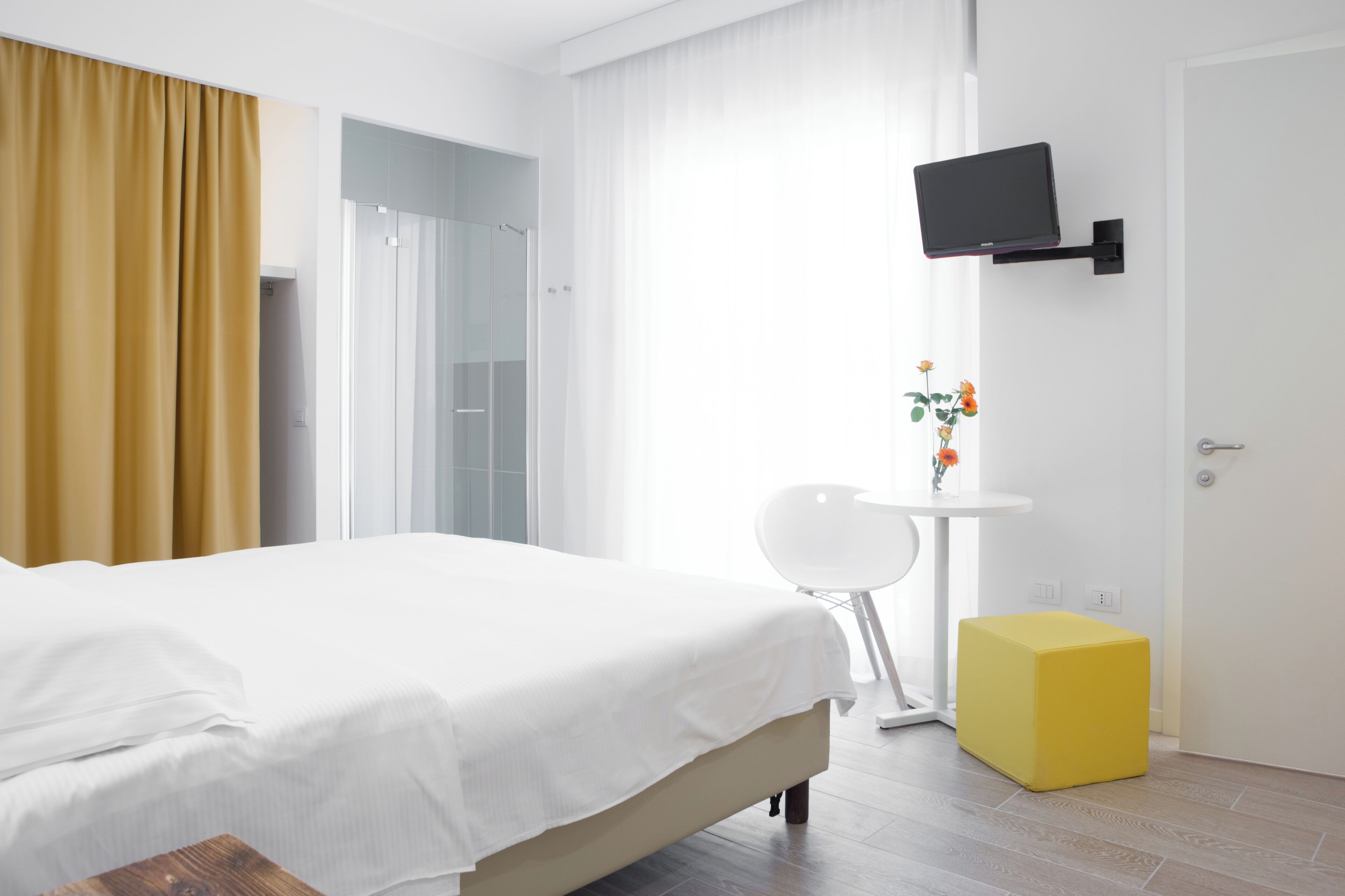 Una camera d'albergo moderna con un letto, una TV a muro, un tavolino con un vaso di fiori e un poggiapiedi giallo.