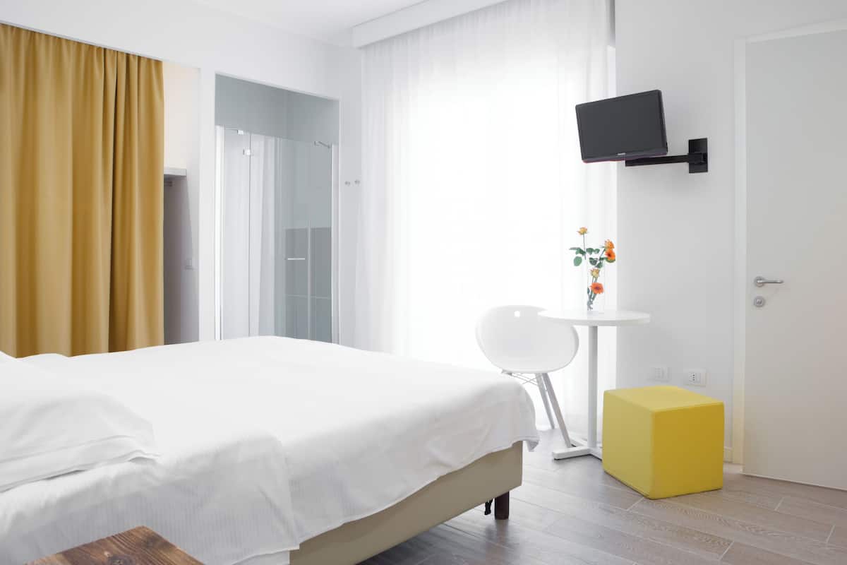 Une chambre d’hôtel moderne avec un lit, un téléviseur fixé au mur, une petite table ornée d’un vase de fleurs et un pouf jaune.
