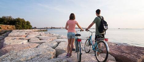 Two people standing on rocky terrain by a body of water, with a distant cityscape in the background.