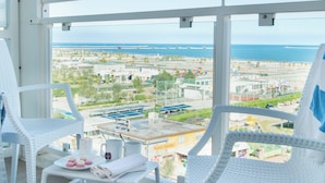 A balcony with white wicker chairs, a small table with a towel and a plate of macarons, and a view of a beach and industrial area. - Smart Hotel Mediterraneo (Chioggia)