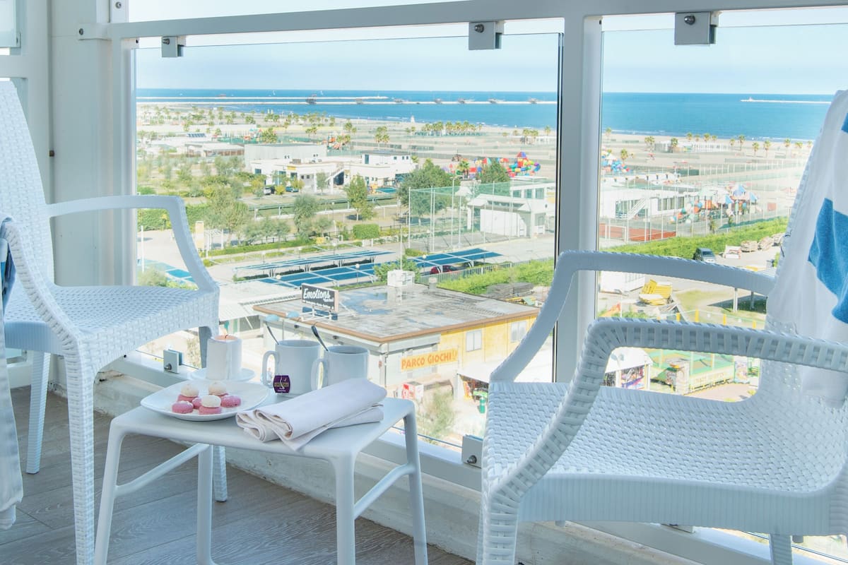 Un balcon avec des fauteuils en osier blancs, une petite table avec une serviette et une assiette de macarons, et une vue sur une plage et une zone industrielle.