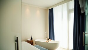 A modern bathroom with a freestanding bathtub, a wooden bench, and a bedside table with a bottle and glasses. - Smart Hotel Mediterraneo (Chioggia)