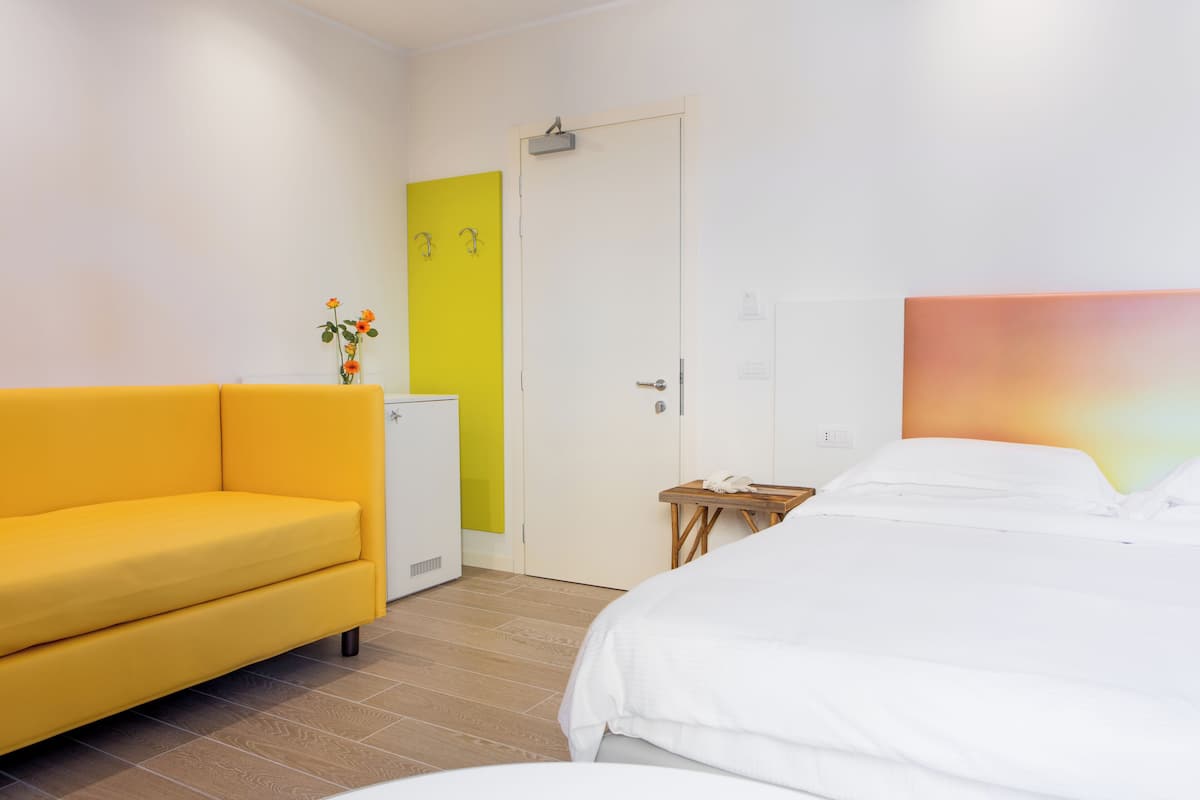 Une chambre d’hôtel avec un canapé jaune, un lit avec une literie blanche, une petite table avec un téléphone et une porte agrémentée d’une touche de jaune.