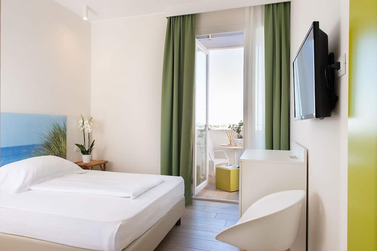 Une chambre d’hôtel avec un lit, un bureau, une chaise, une télévision et une vue sur la mer par la fenêtre.
