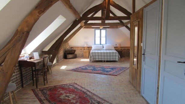 A loft bedroom with a sloped ceiling, exposed wooden beams, a bed, a desk, and a large rug. - A l'Ecole Buissonnière (Honfleur)