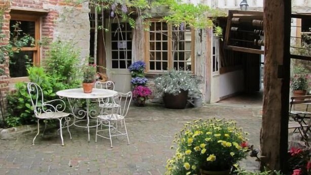 A courtyard with a brick wall, a wooden pergola, a round table with chairs, and potted plants. - A l'Ecole Buissonnière (Honfleur)